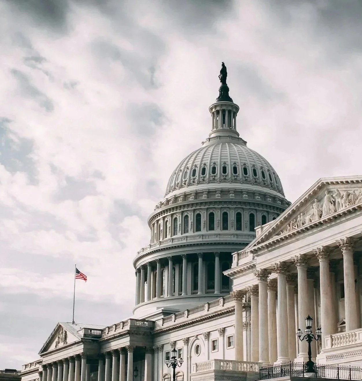 The United States Capitol building in Washington, D.C., featuring its large dome, columns, and a flagpole with an American flag, under a cloudy sky.