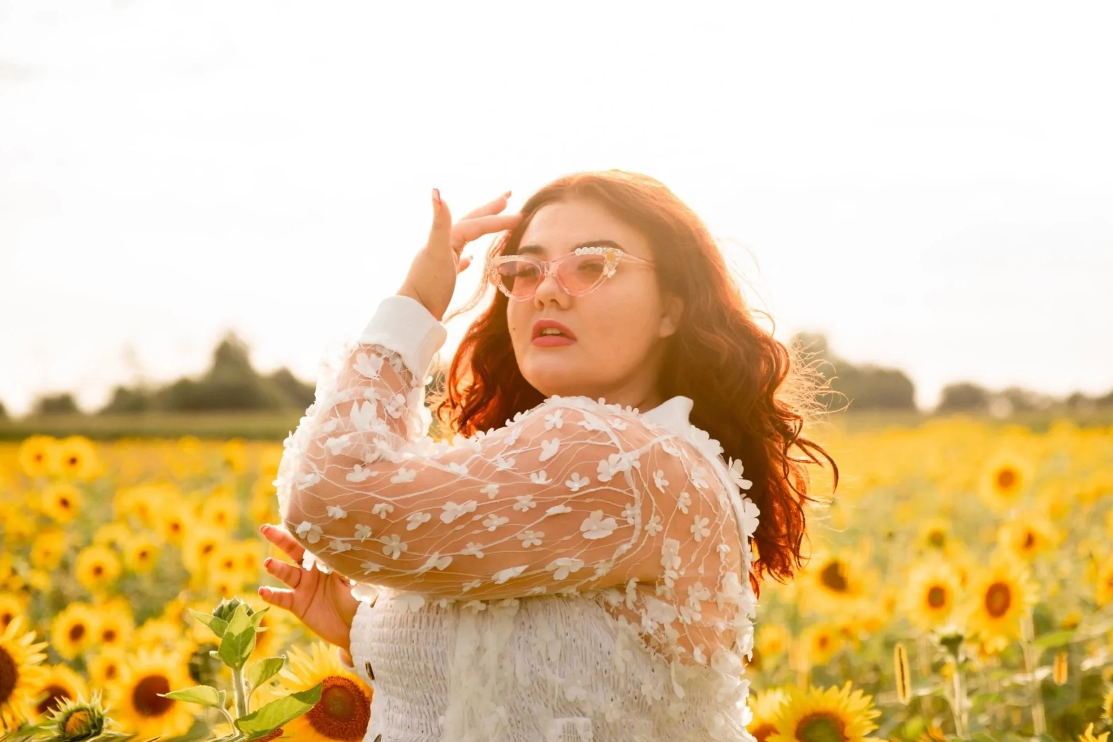 A woman with reddish curly hair wearing a sheer white floral dress and pink sunglasses stands in a sunflower field during sunset.