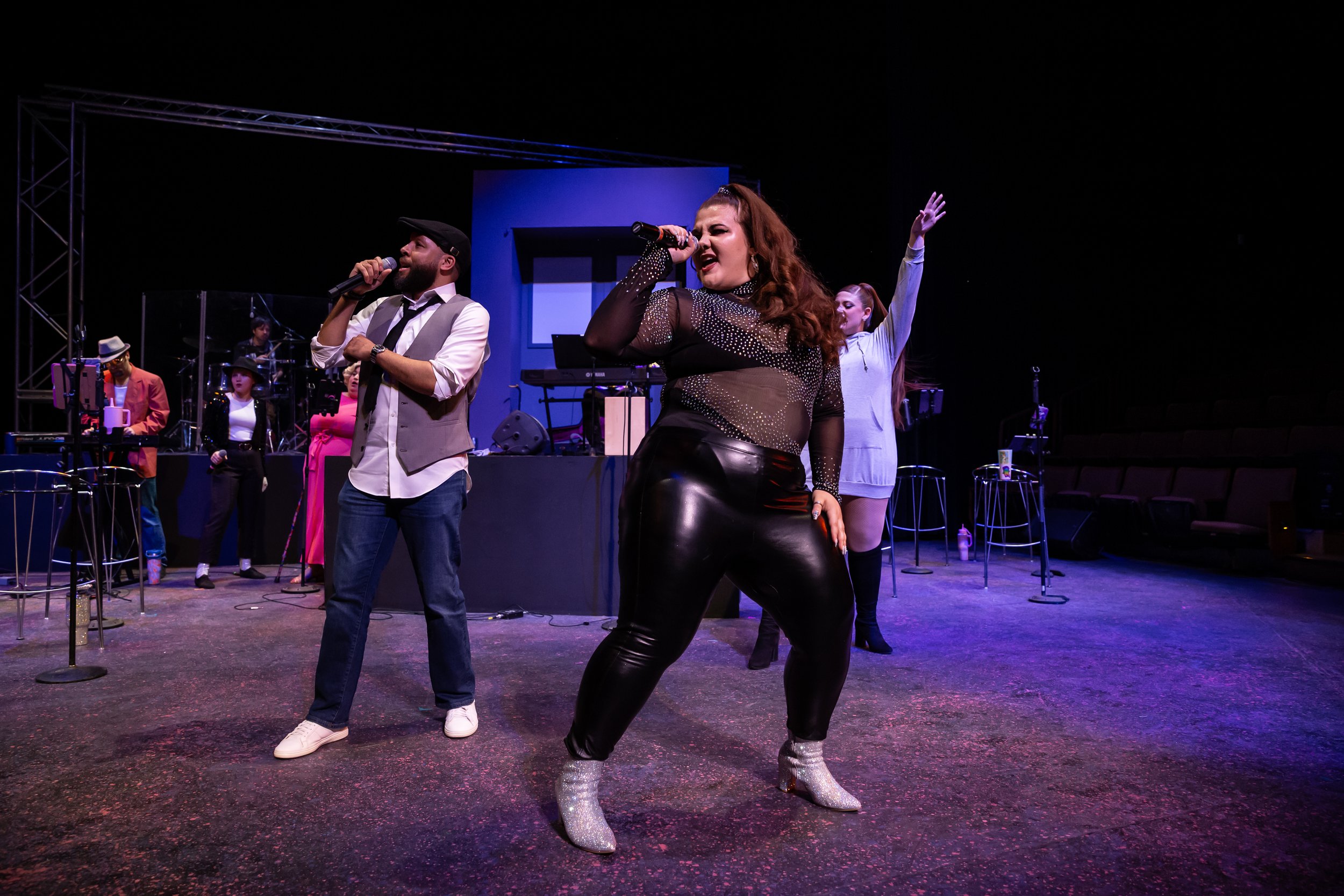 Group of performers on stage, singing and dancing with musical instruments and microphones, colorful stage lighting, and a dark background.