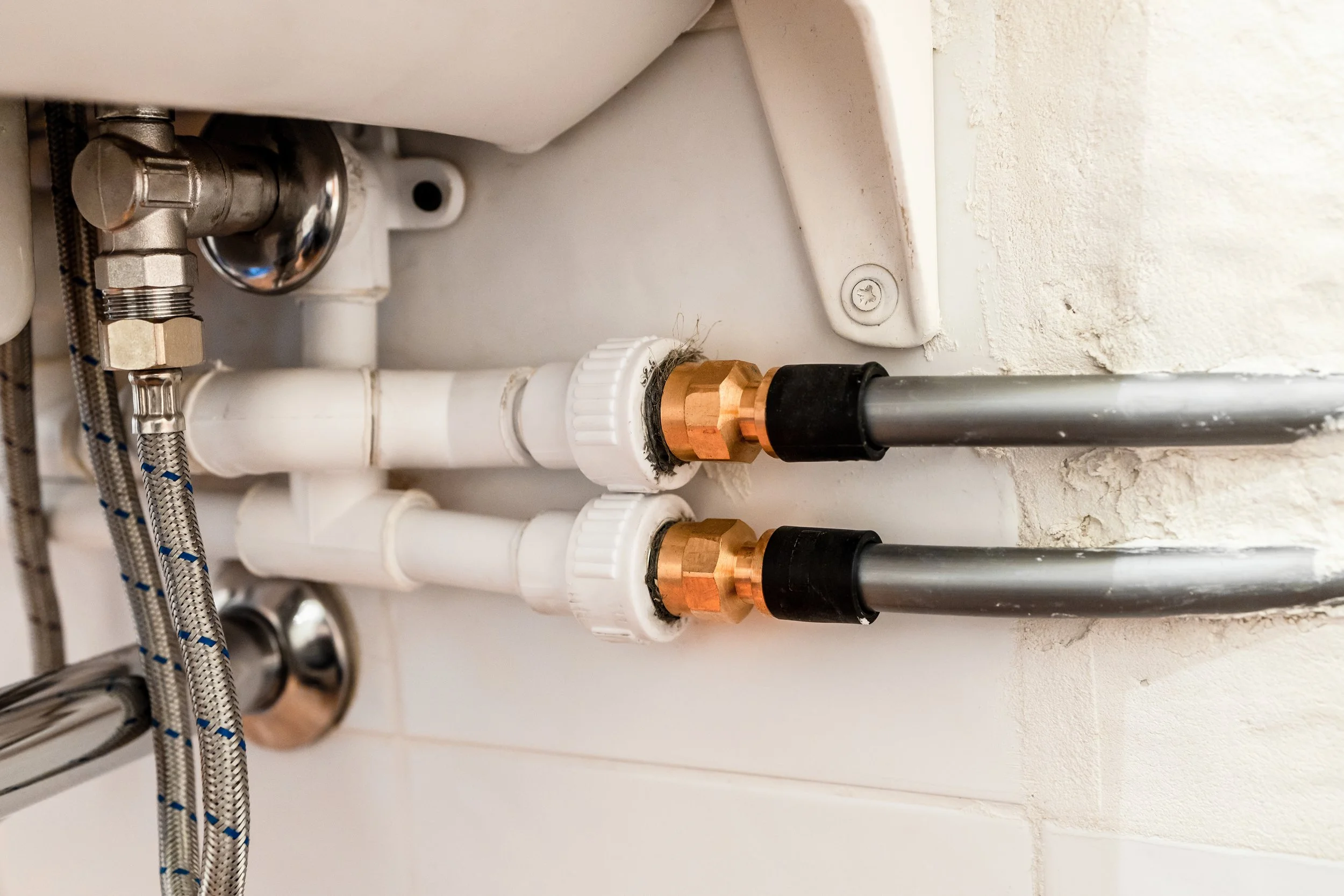 Close-up of plumbing pipes and fittings under a sink, including white plastic pipes, brass connectors, and metal supply lines.