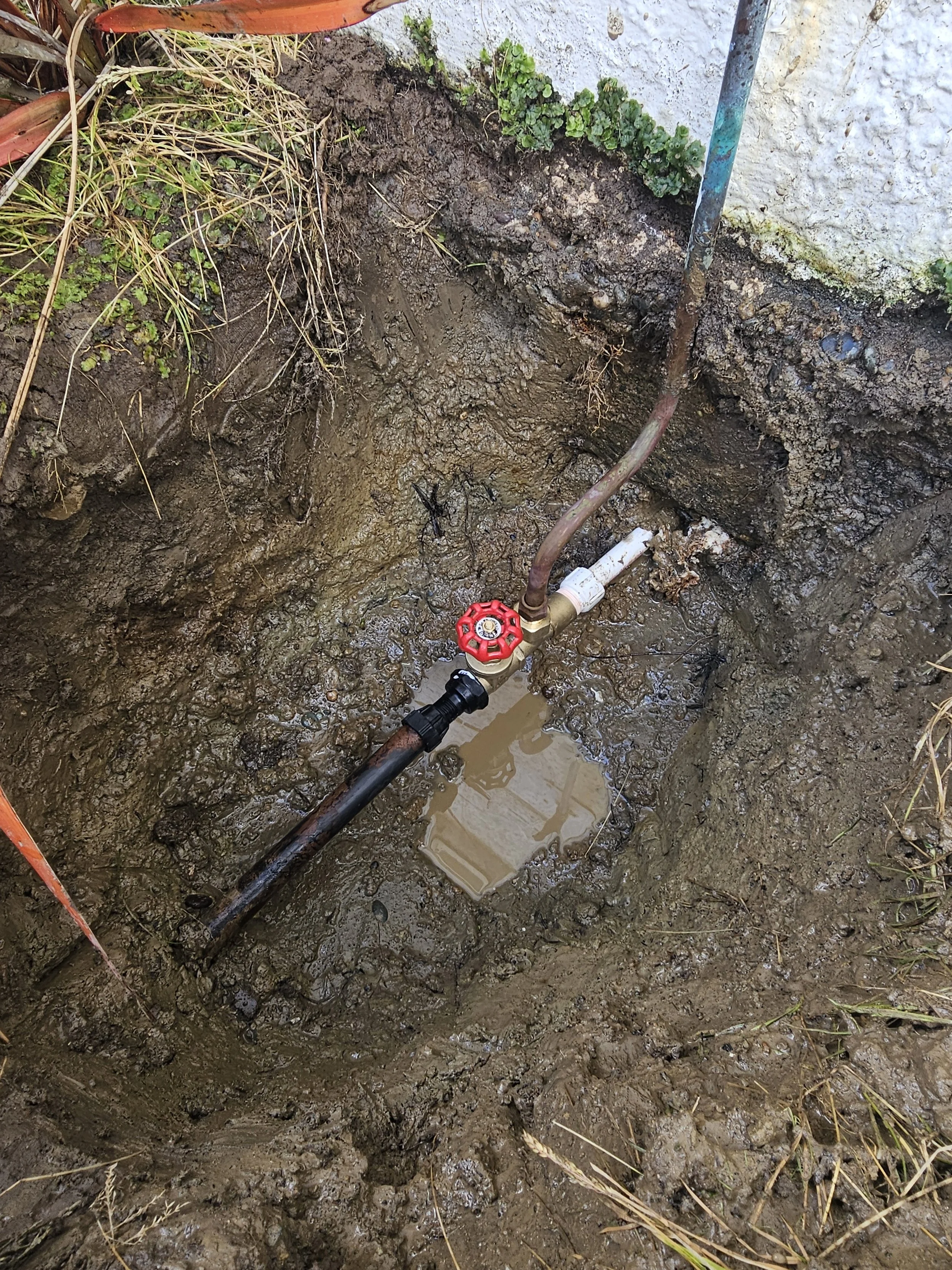 A close-up view of a small excavated hole in the ground with a water pipe and a valve with a red handle, possibly for plumbing or irrigation, with soil and some grass around the excavation.
