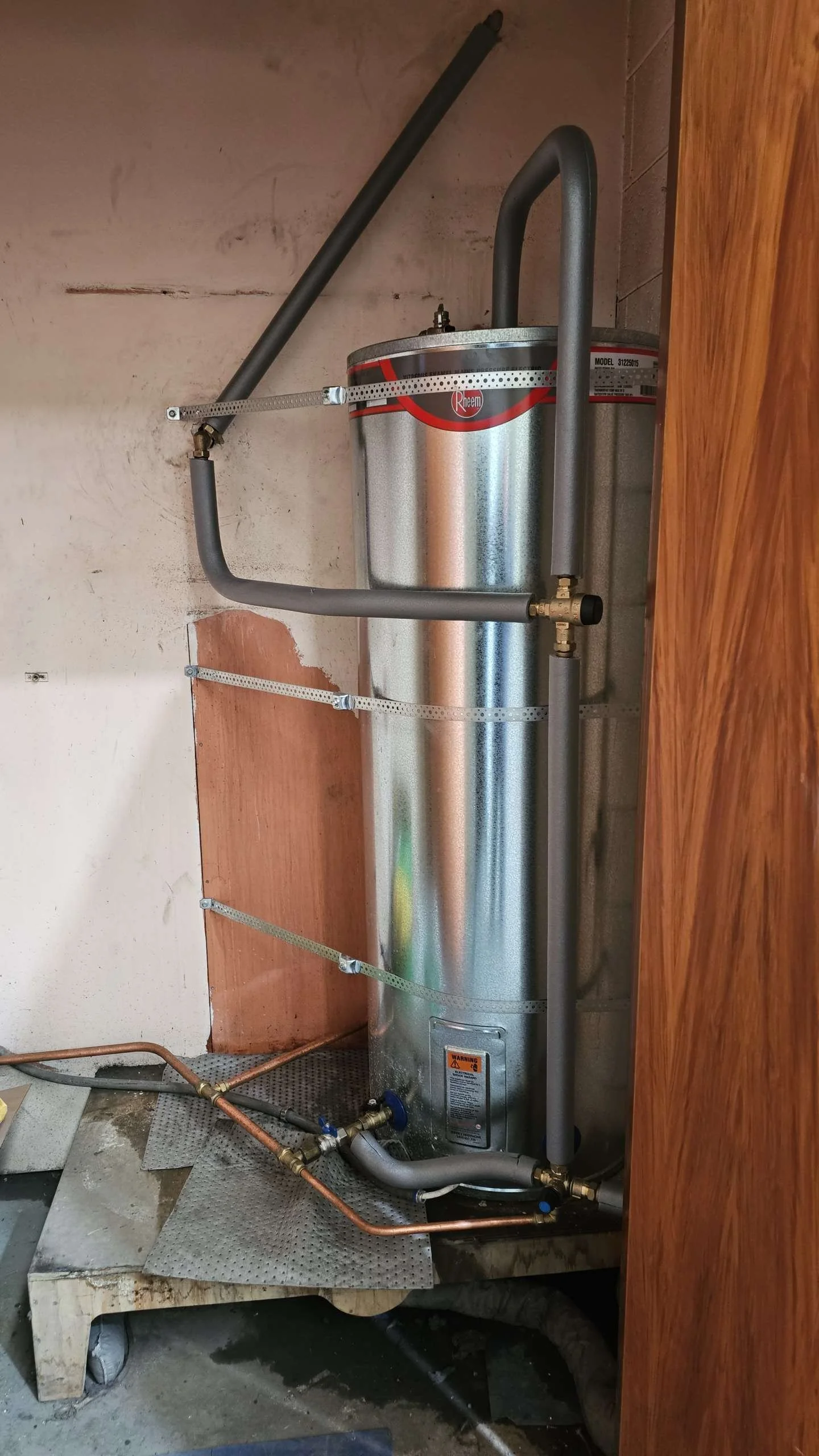 A water heater installed in a closet with visible pipes and wood paneling.