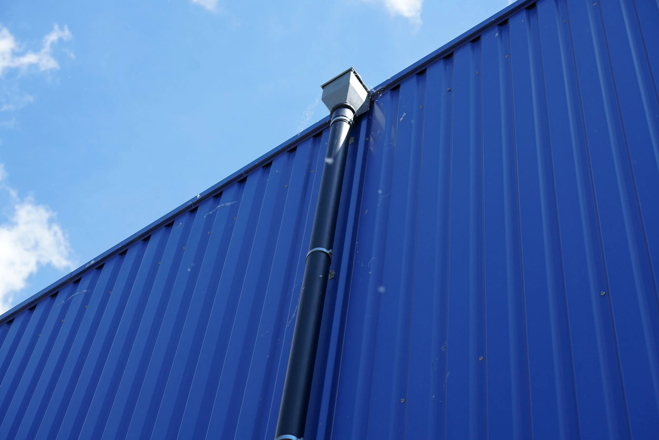 Close-up of a black pipe running down the blue metal wall of a building, with the sky and some clouds in the background.