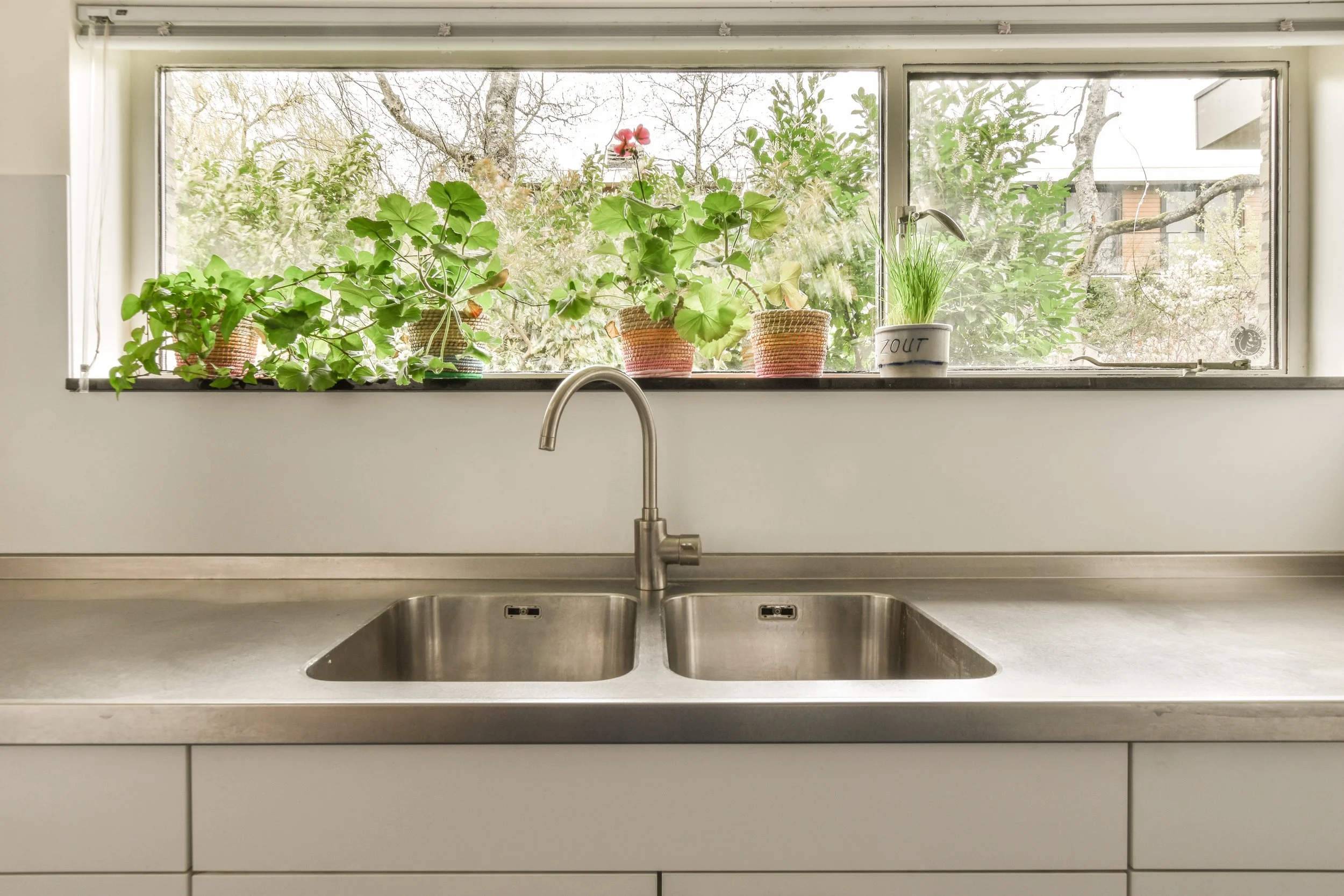 Kitchen sink with stainless steel double basin and a faucet, set beneath a large window with potted plants on the windowsill and a view of trees outside.