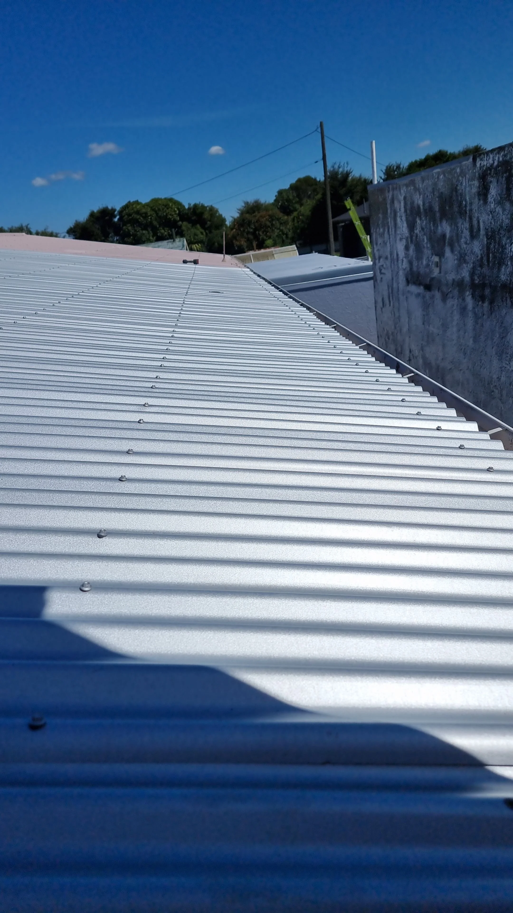 Close-up of a metal corrugated roof under a clear blue sky with some small clouds, trees, and utility poles in the background.