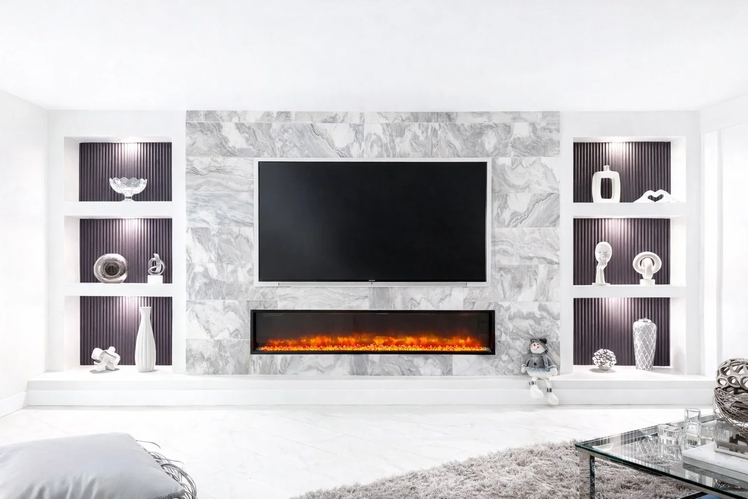 Modern living room with a mounted flat-screen TV above an electric fireplace with orange flames, surrounded by white marble wall, flanked by built-in bookshelves with decorative white vases and sculptures, white walls, a glass coffee table, and a gray area rug.