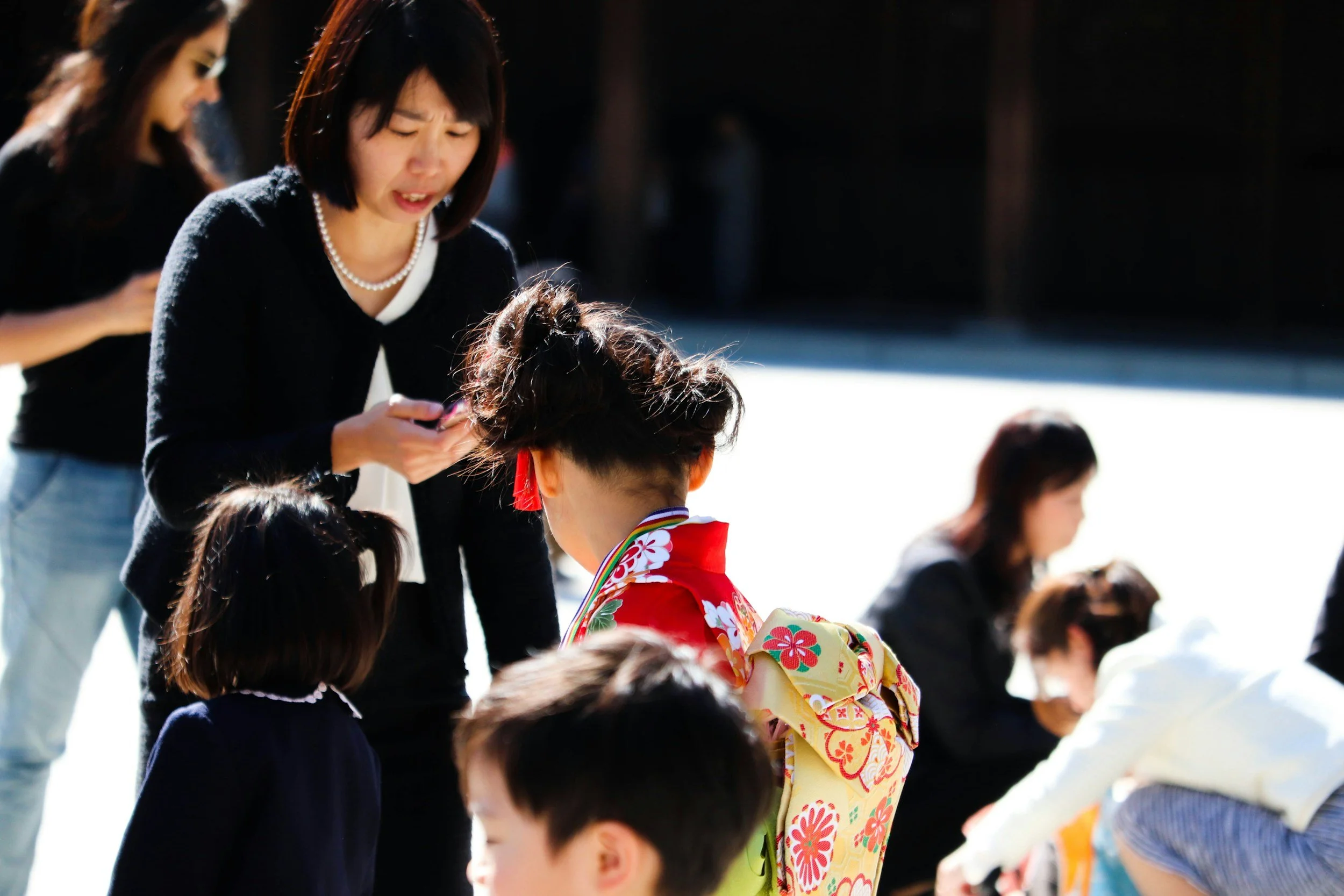 A young girl in a colorful traditional Japanese kimono with a floral pattern and a red obi, standing among a group of children, dressed in dark clothing, with a woman in the background and others around, outdoors on a bright day.