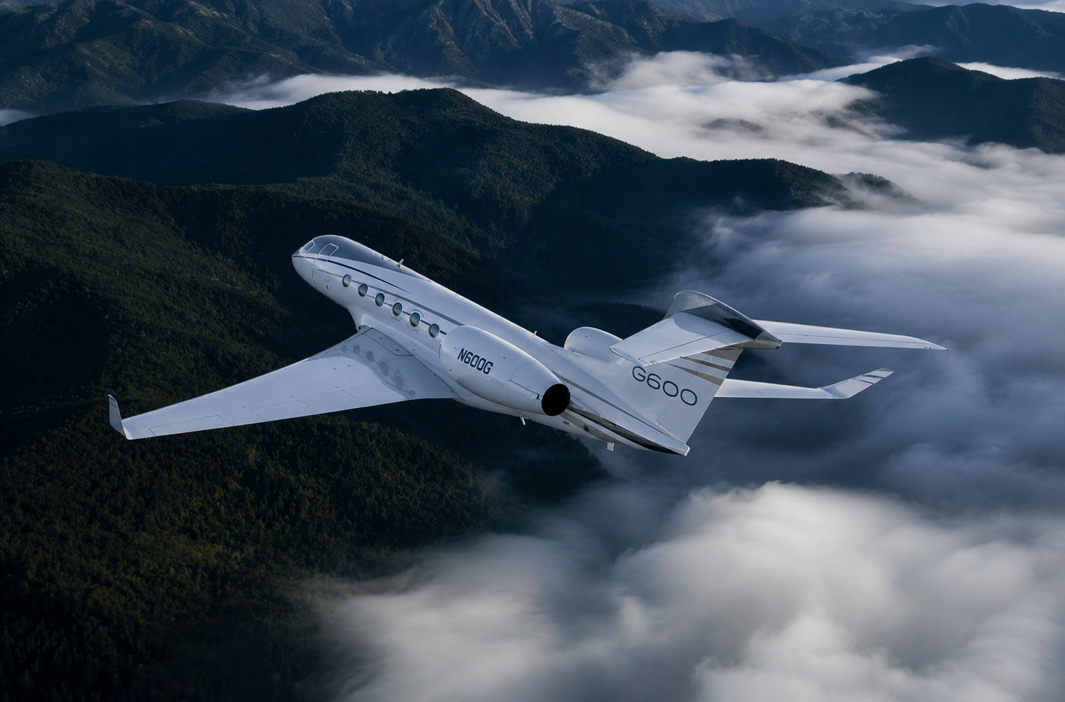 A white G600 jet airplane flying above green mountain ridges surrounded by clouds and fog.