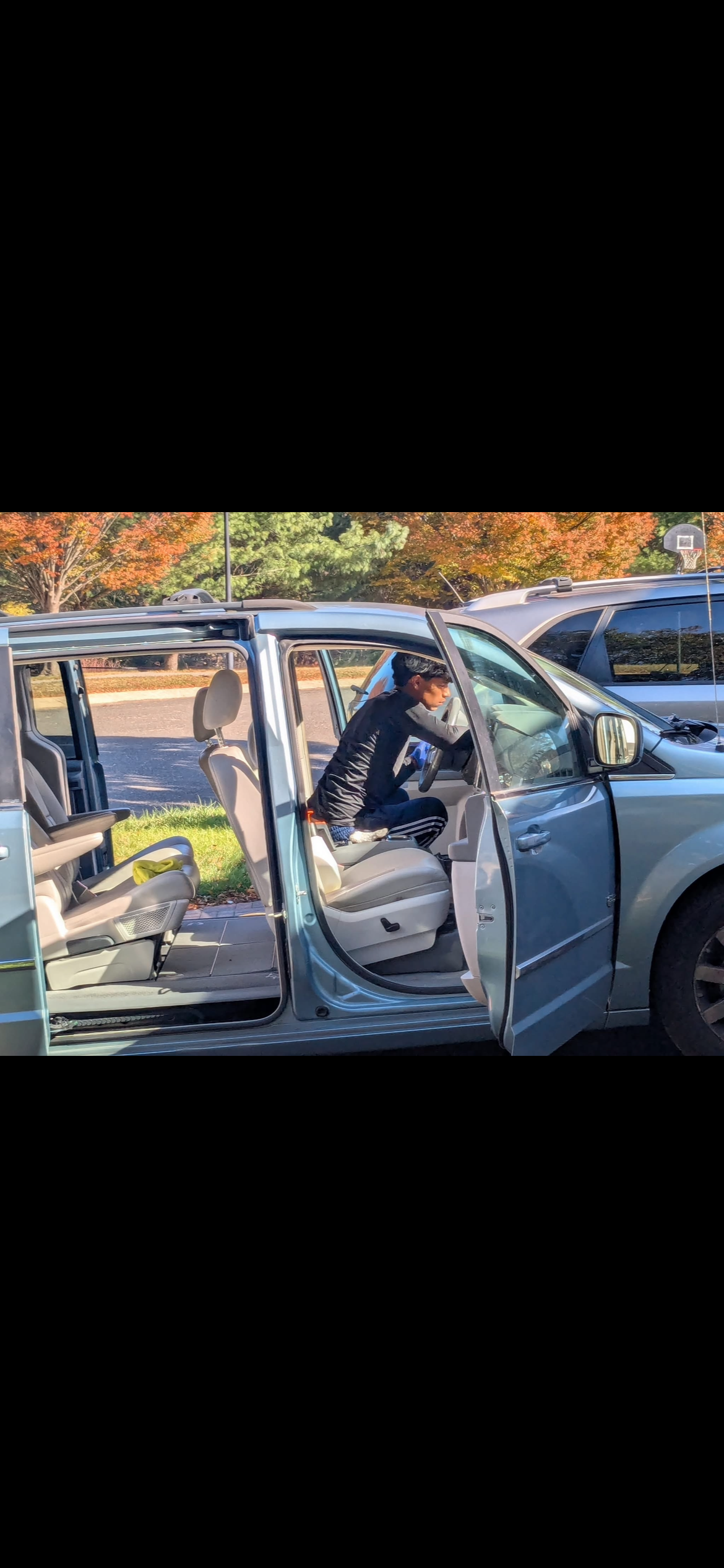 A person sitting in the driver's seat of a blue minivan, looking towards the steering wheel, with the passenger door open, parked in a lot with trees in autumn foliage in the background.