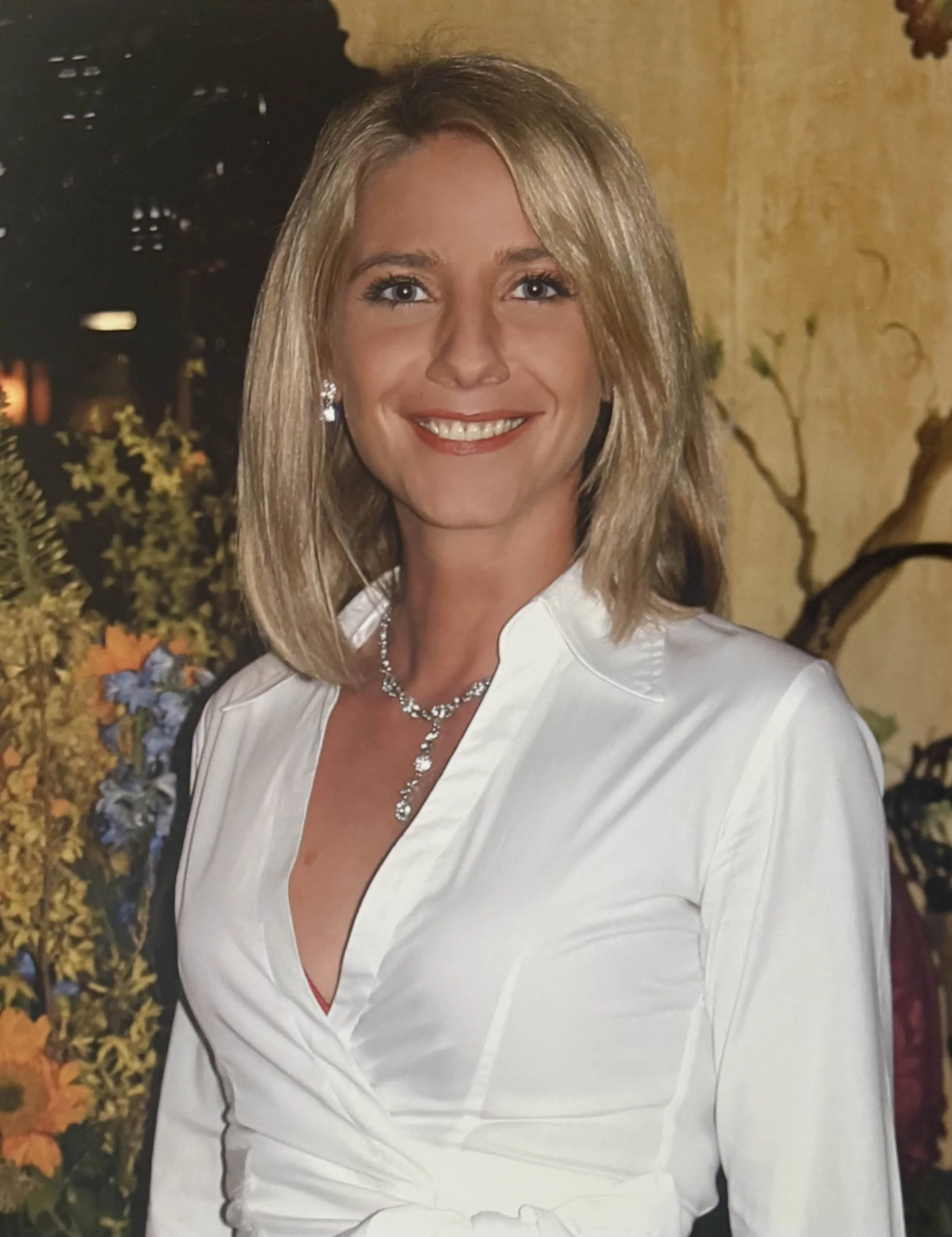 A woman with blonde hair smiling, wearing a white blouse and jewelry.