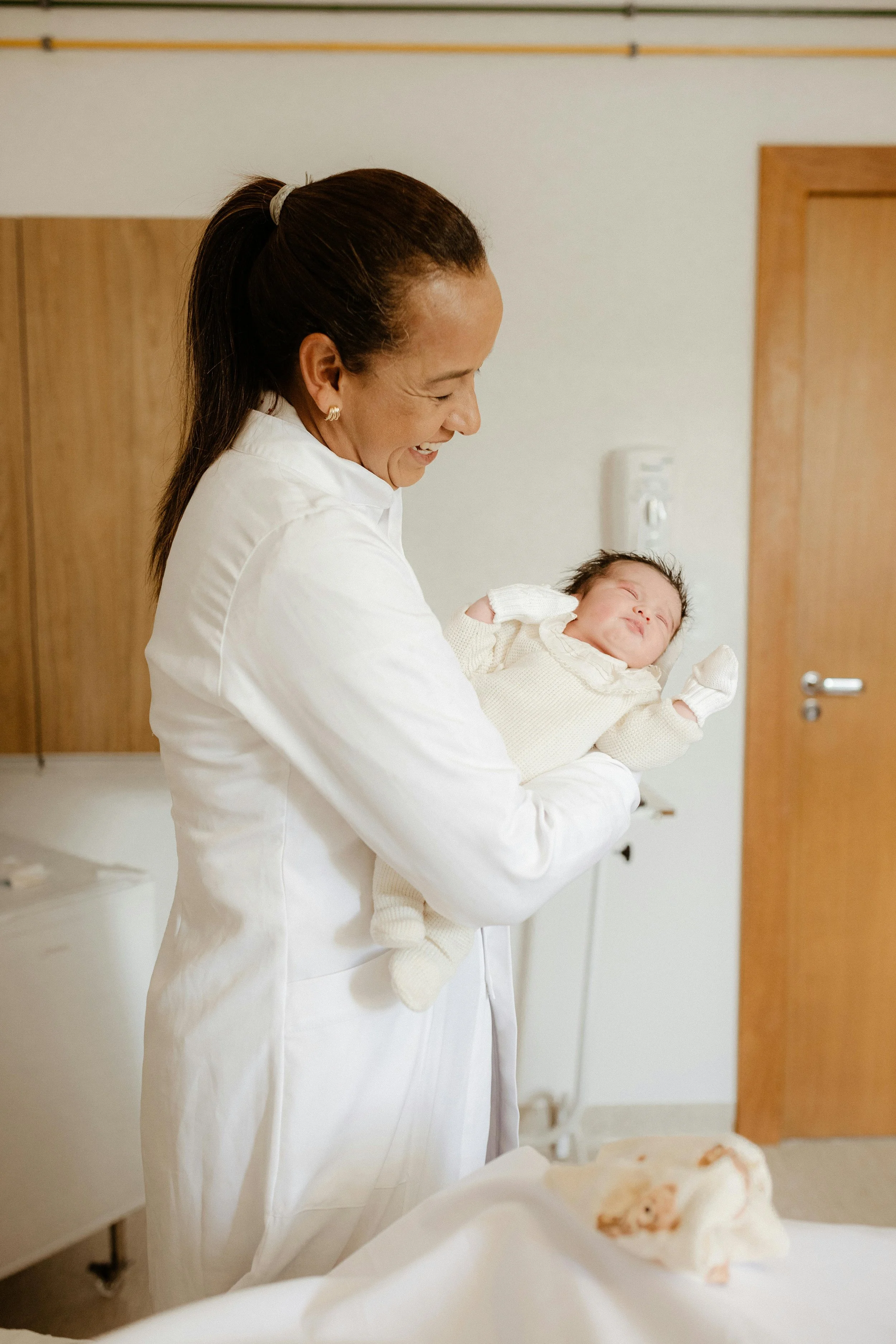 A woman in a white coat holding a happy newborn baby dressed in cream-colored clothing in a hospital room.