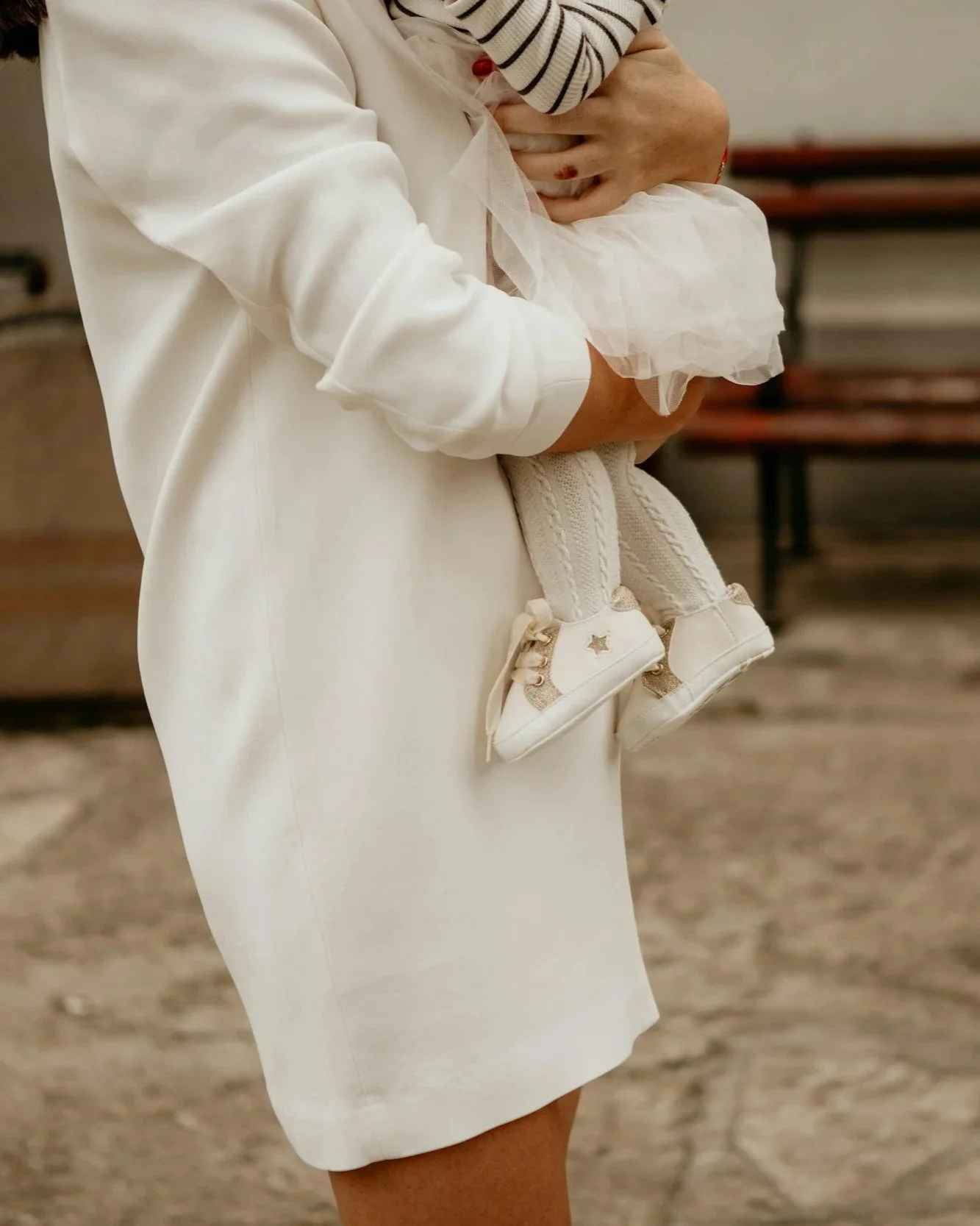 Close-up of a woman holding a small child, both dressed in white, with the woman wearing a white blazer and the child in white shoes and tights, set against a blurred background.