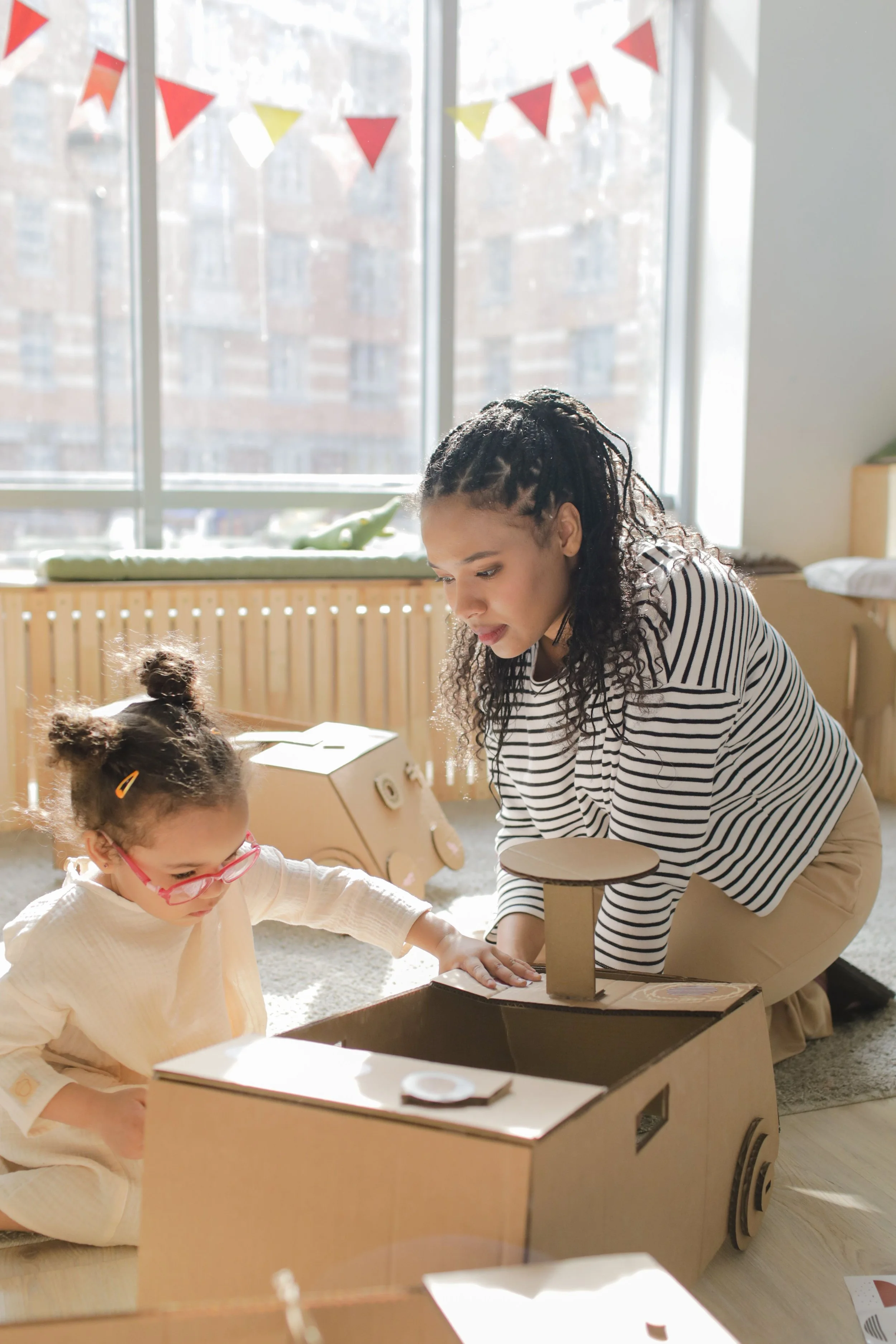 A woman and a young girl are playing with a cardboard pretend car indoors. The woman has curly hair and is wearing a black and white striped top. The girl has her hair in pigtails, glasses, and is wearing a cream-colored outfit. Sunlight streams in through large windows decorated with red and yellow triangular flags.