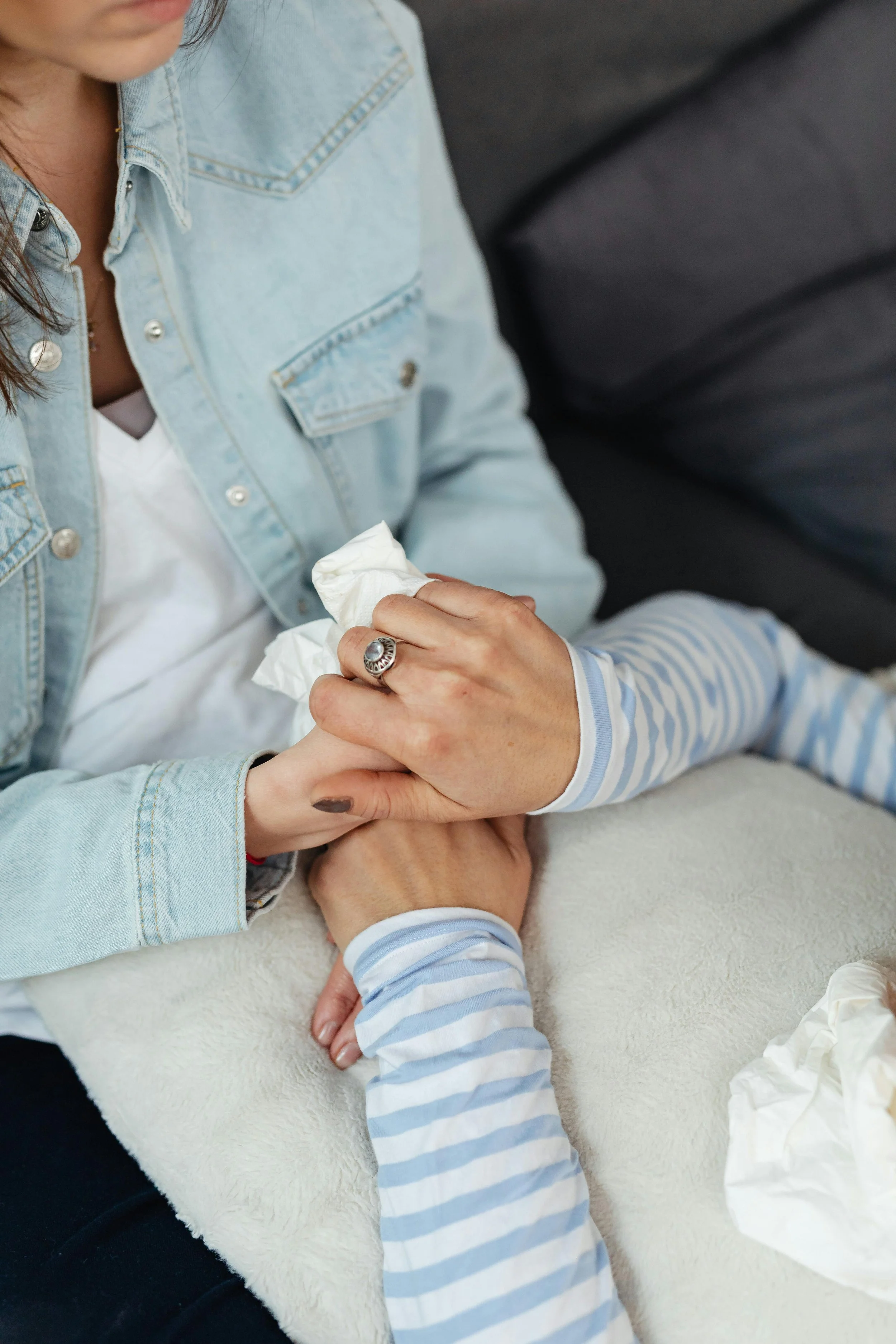 Close-up of two hands, one adult and one child, holding each other in a comforting manner. The adult is wearing a denim jacket and a ring, while the child is wearing a striped long-sleeve shirt. The background shows a soft surface with crumpled tissues nearby.