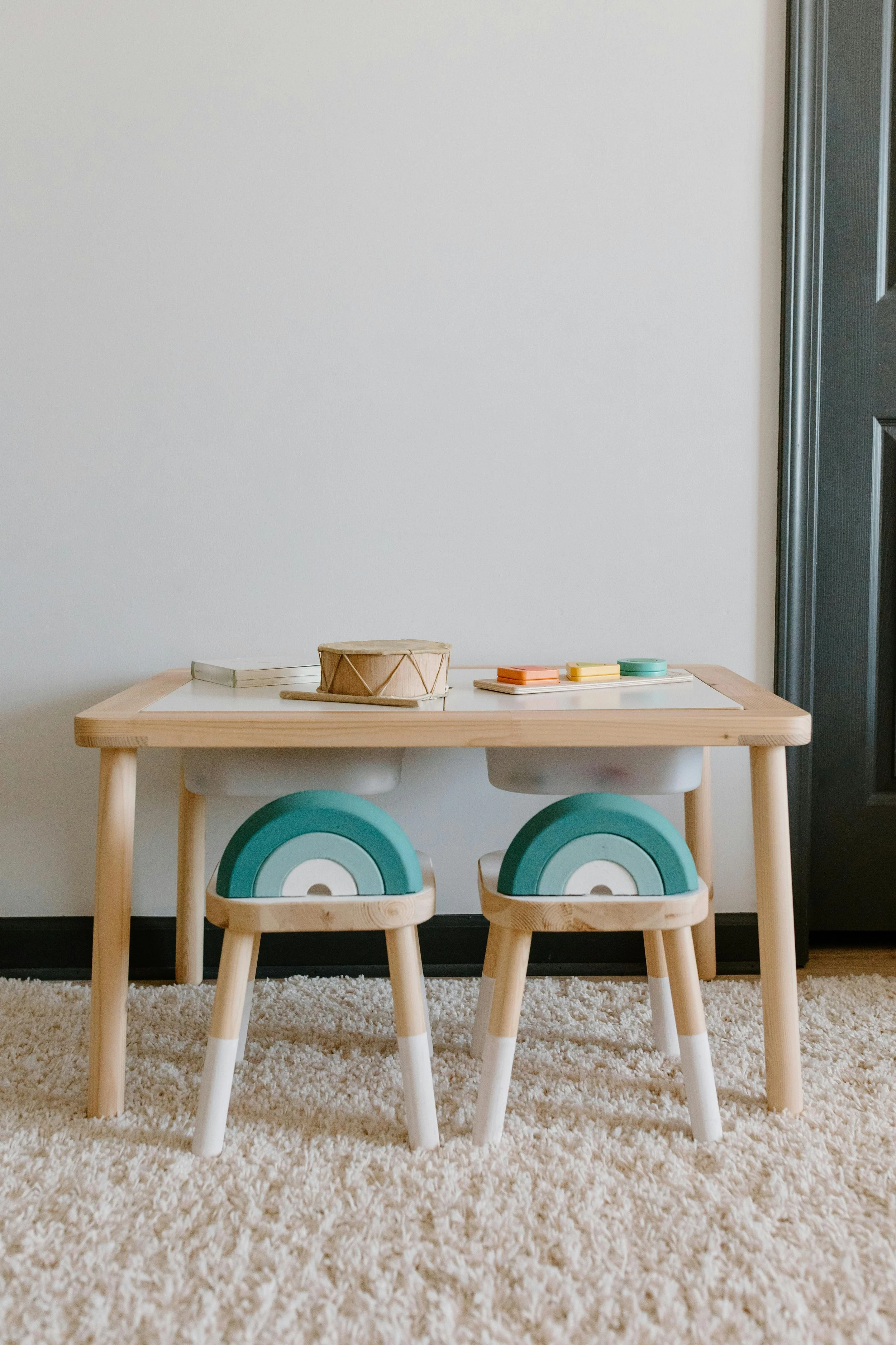 A small wooden table with two matching wooden stools beneath it. On the table, there is a small drum, some pieces of paper, and some colorful round and rectangular objects, possibly toys or educational materials. The setting appears to be a child's play area or classroom, with a beige carpet and plain wall background.
