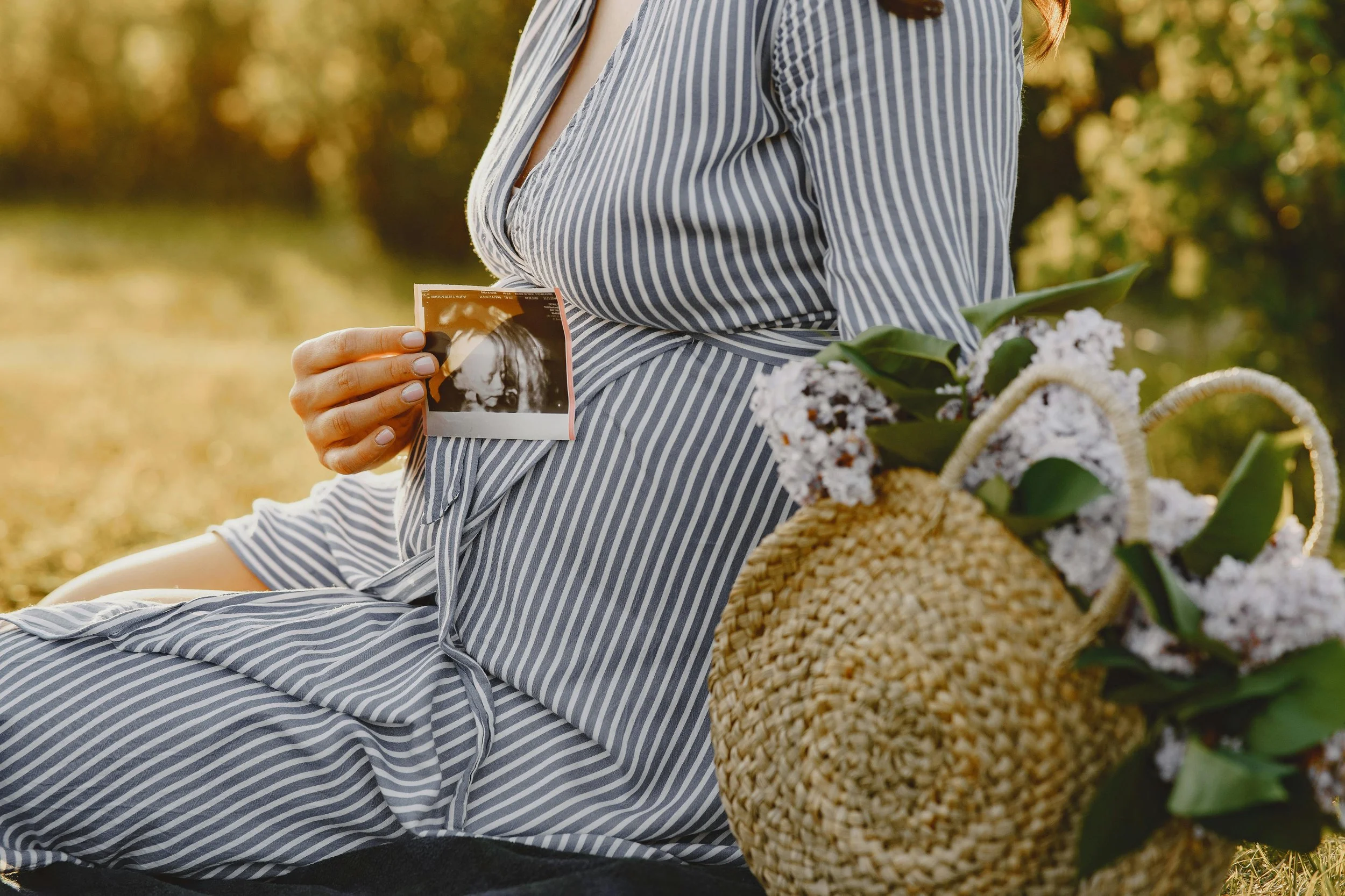 A pregnant woman sitting outdoors holding an ultrasound picture, with a woven bag filled with white flowers next to her.