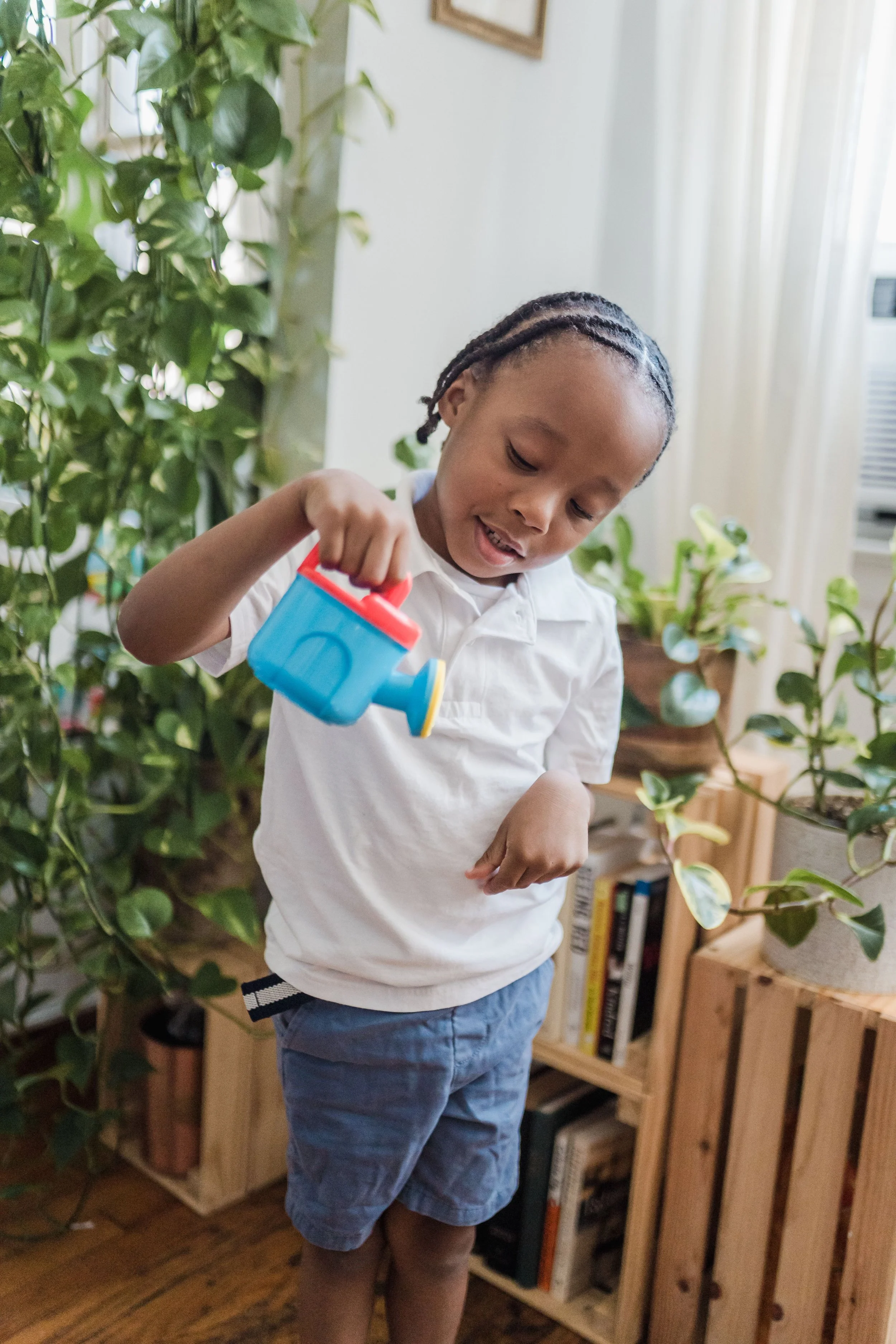 A young boy with braided hair wearing a white shirt and blue shorts, holding a toy watering can in an indoor setting with green plants and a bookshelf in the background.