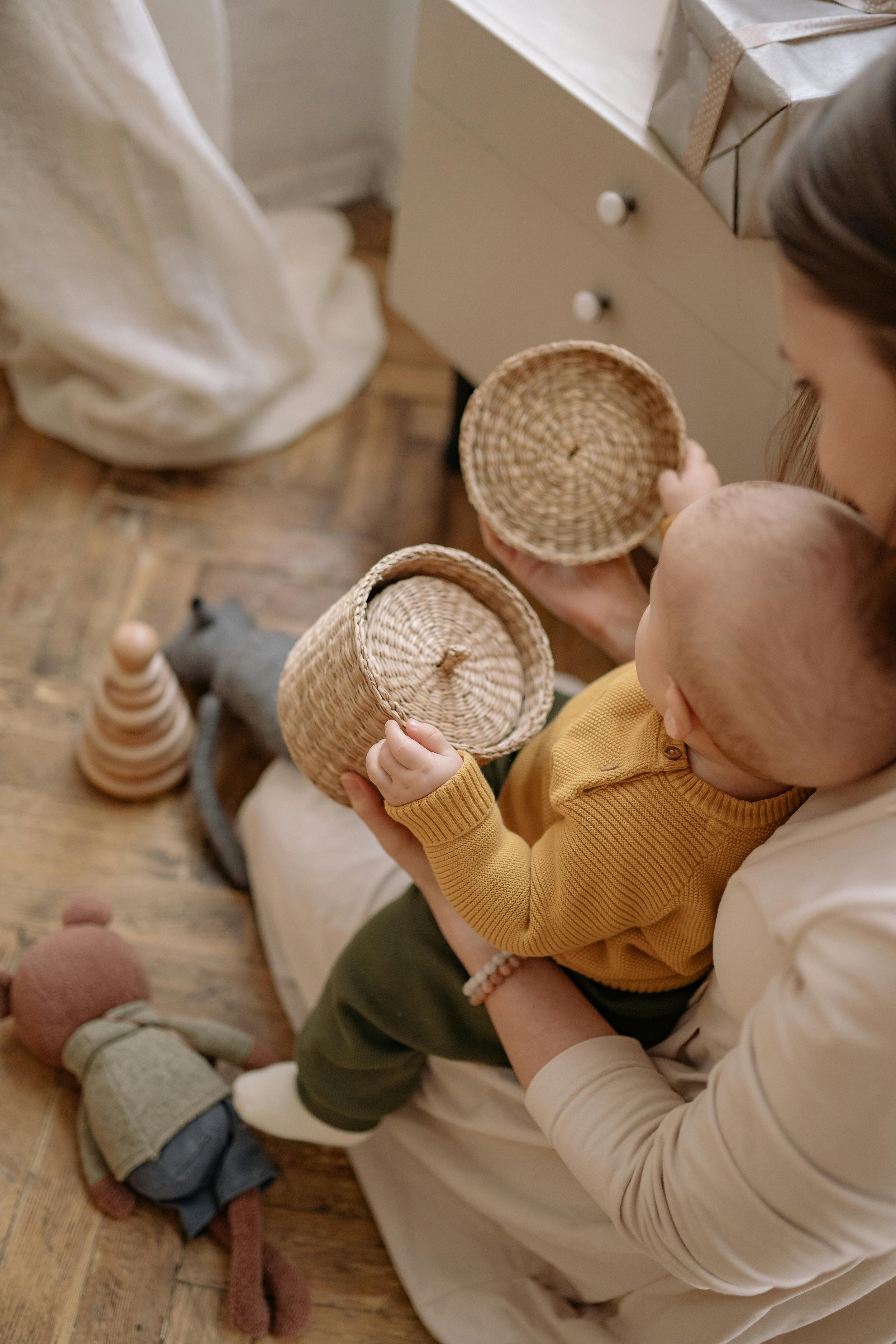 A woman holding a baby on her lap, both holding woven baskets, in a cozy room with wooden flooring and toys nearby.