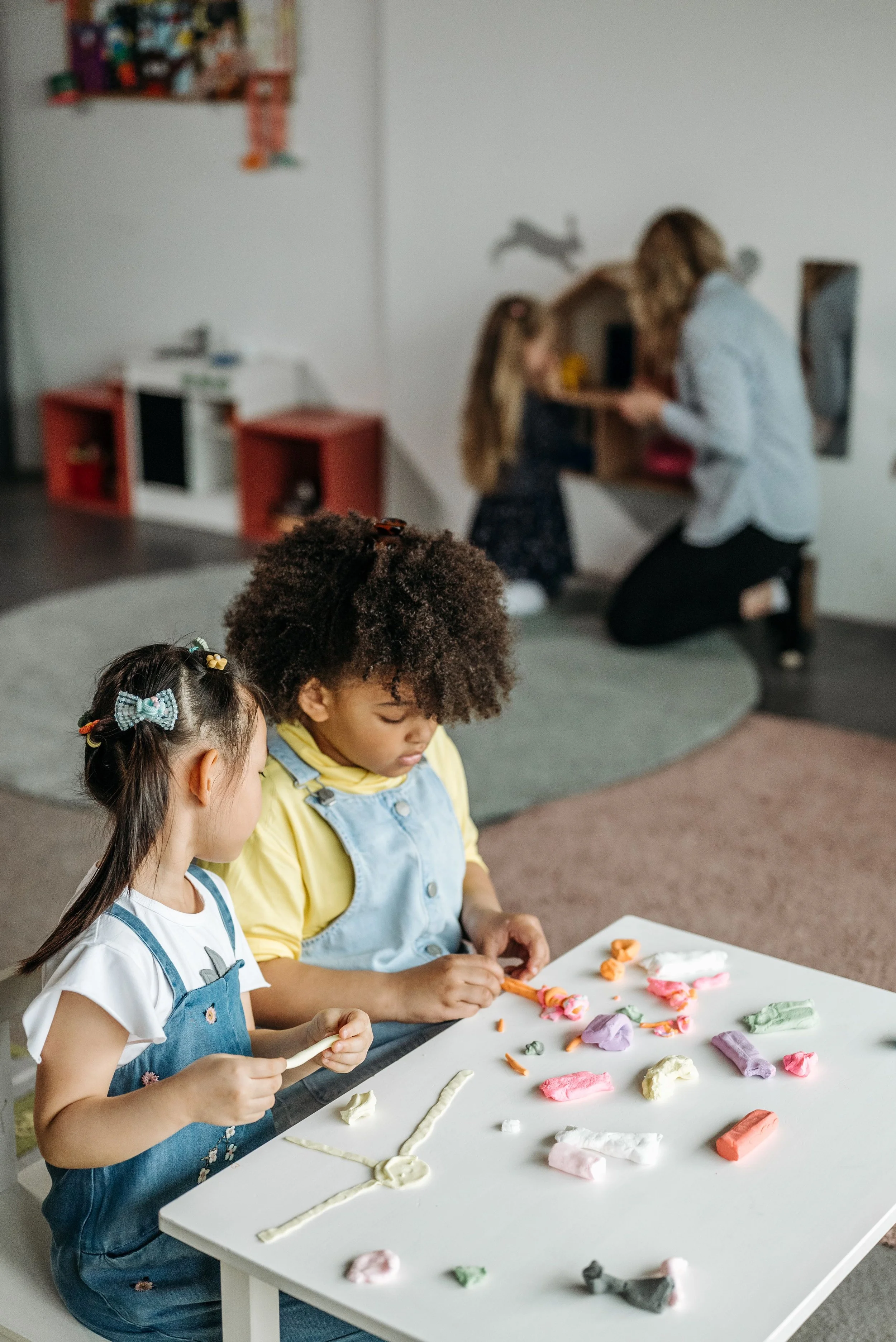 Children creating colorful clay sculptures on a white table in a classroom setting with a woman helping in the background.