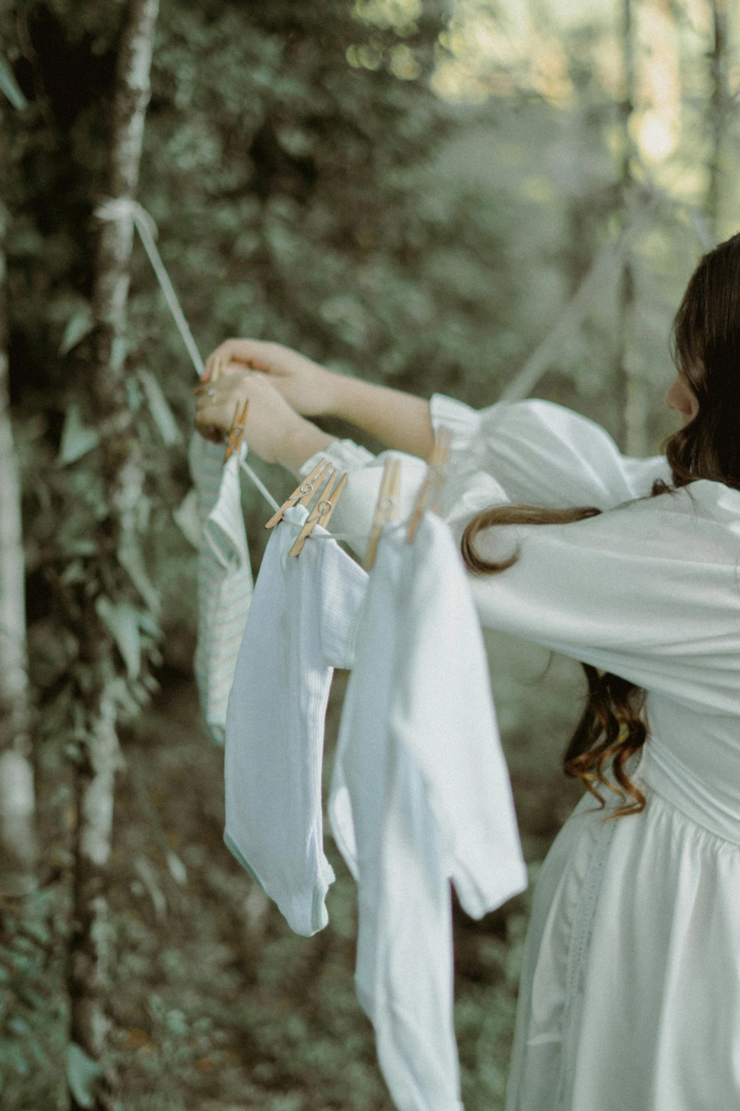 A woman with long dark hair hanging over her shoulder, wearing a white dress, hanging baby clothes on a clothesline in a forested area.