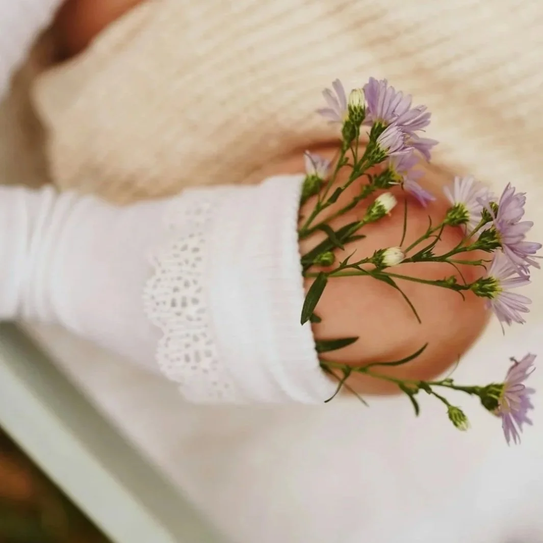 A baby's hand holding a sprig of purple daisies, wearing a white lace sleeve.