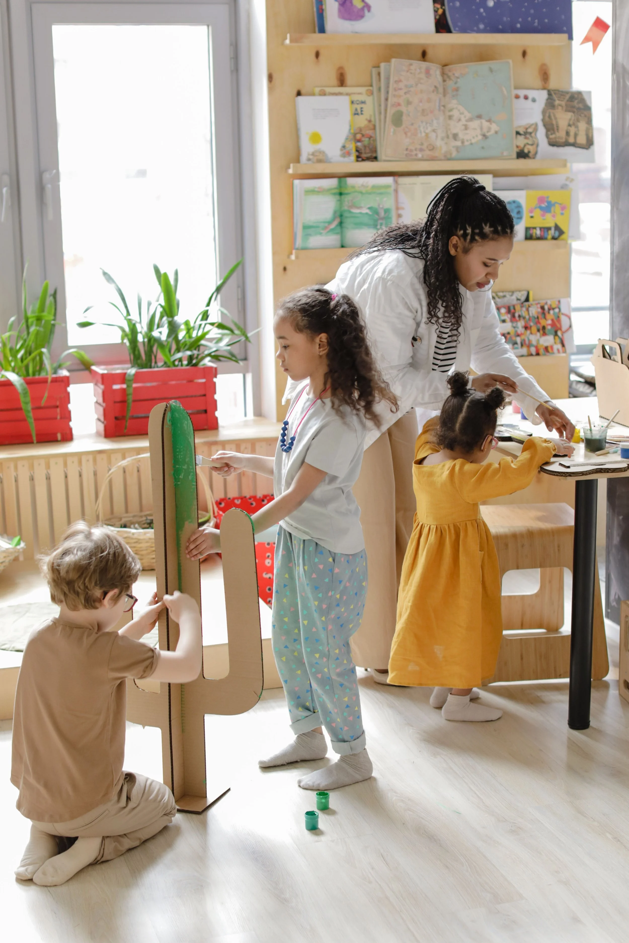 Children and a teacher engaging in a painting activity in a classroom, with some children sitting and others standing around a table.