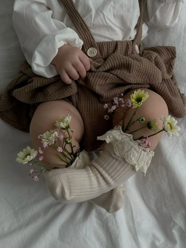 A young child sitting on a bed with their legs spread, wearing a white shirt, a brown skirt, and white knee-high socks with lace trim. The child's knees are decorated with small flowers.