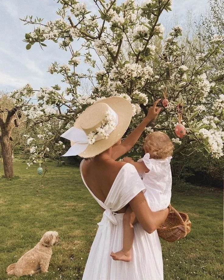 Woman in a white dress and large sun hat holding a young child, both reaching for hanging Easter eggs on a blooming tree in a grassy park. A small, tan dog sits on the grass watching them.