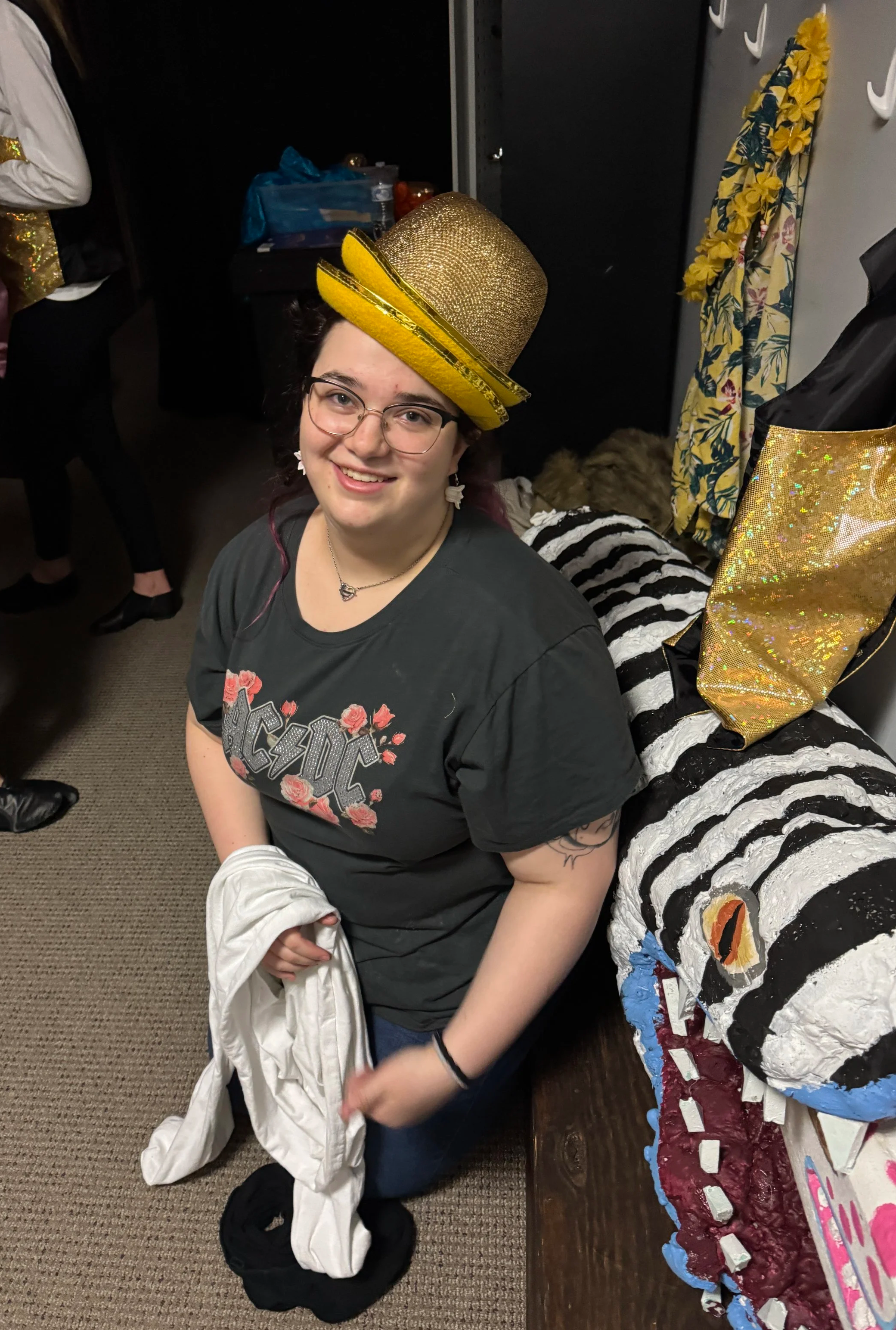 A woman wearing a gold top hat with yellow band, glasses, black AC/DC t-shirt, holding a white jacket, smiling, kneeling on a carpeted floor at a party with a decorated cake in front of her.
