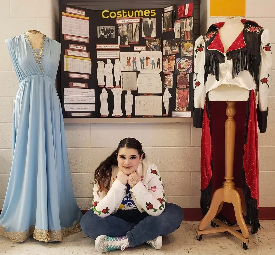 A girl sitting on the floor with her chin resting on her hands, smiling, surrounded by costume displays and a bulletin board about costumes.