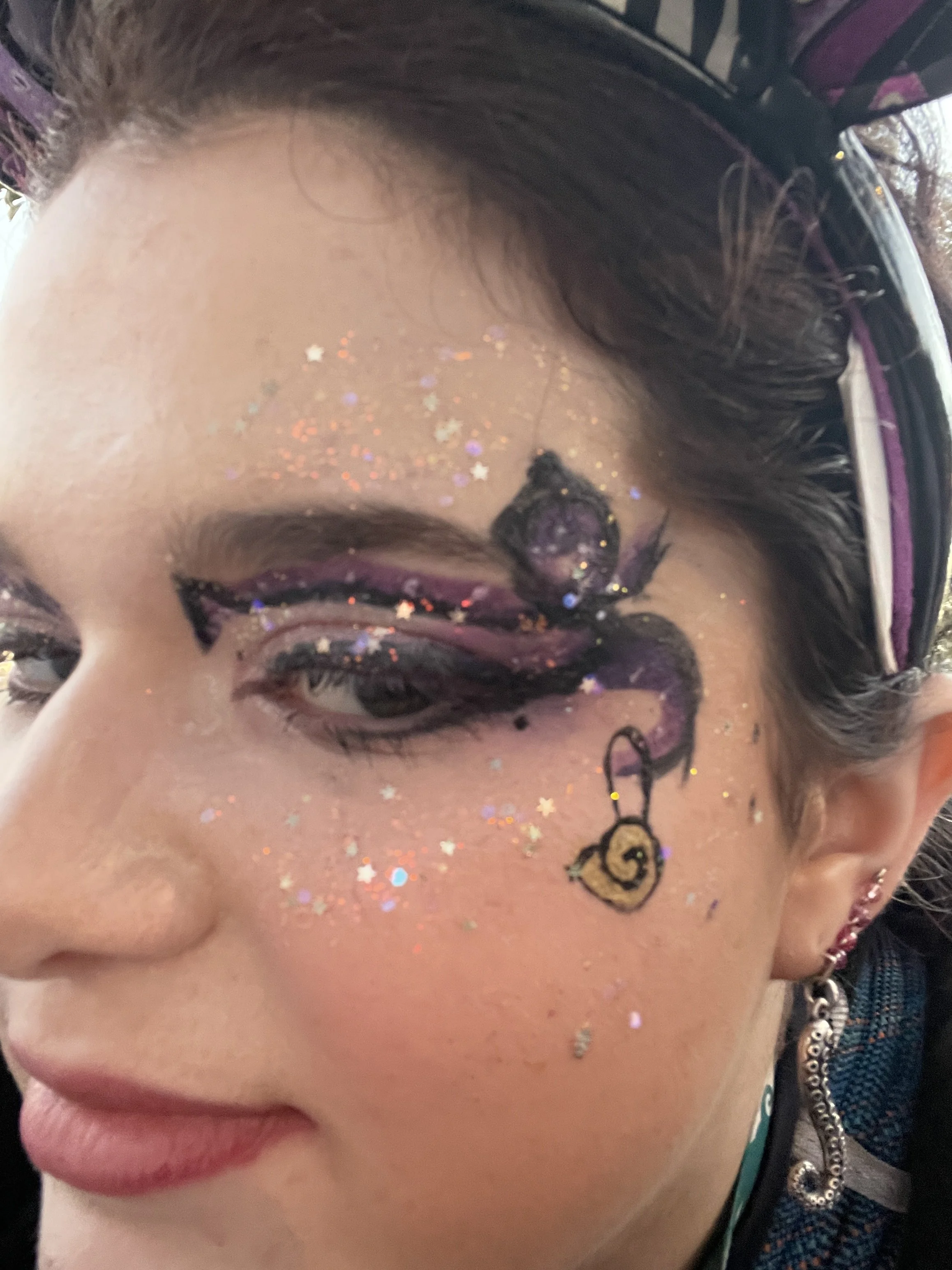 A close-up of a woman's face with elaborate makeup and glitter, including a design around her eye with black and purple accents, star-shaped glitter, and a small cartoon snail charm drawing below her eye.