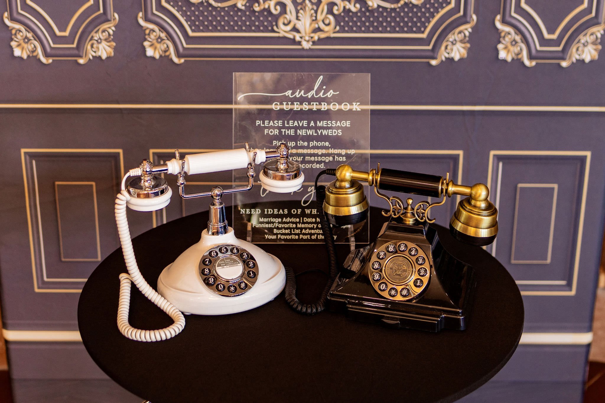 Two vintage rotary dial telephones, one white and one black with gold accents, are placed on a black table. Behind them is a transparent sign on a purple and gold decor background that reads 'Audio Guestbook' and instructions for leaving a message for newlyweds.