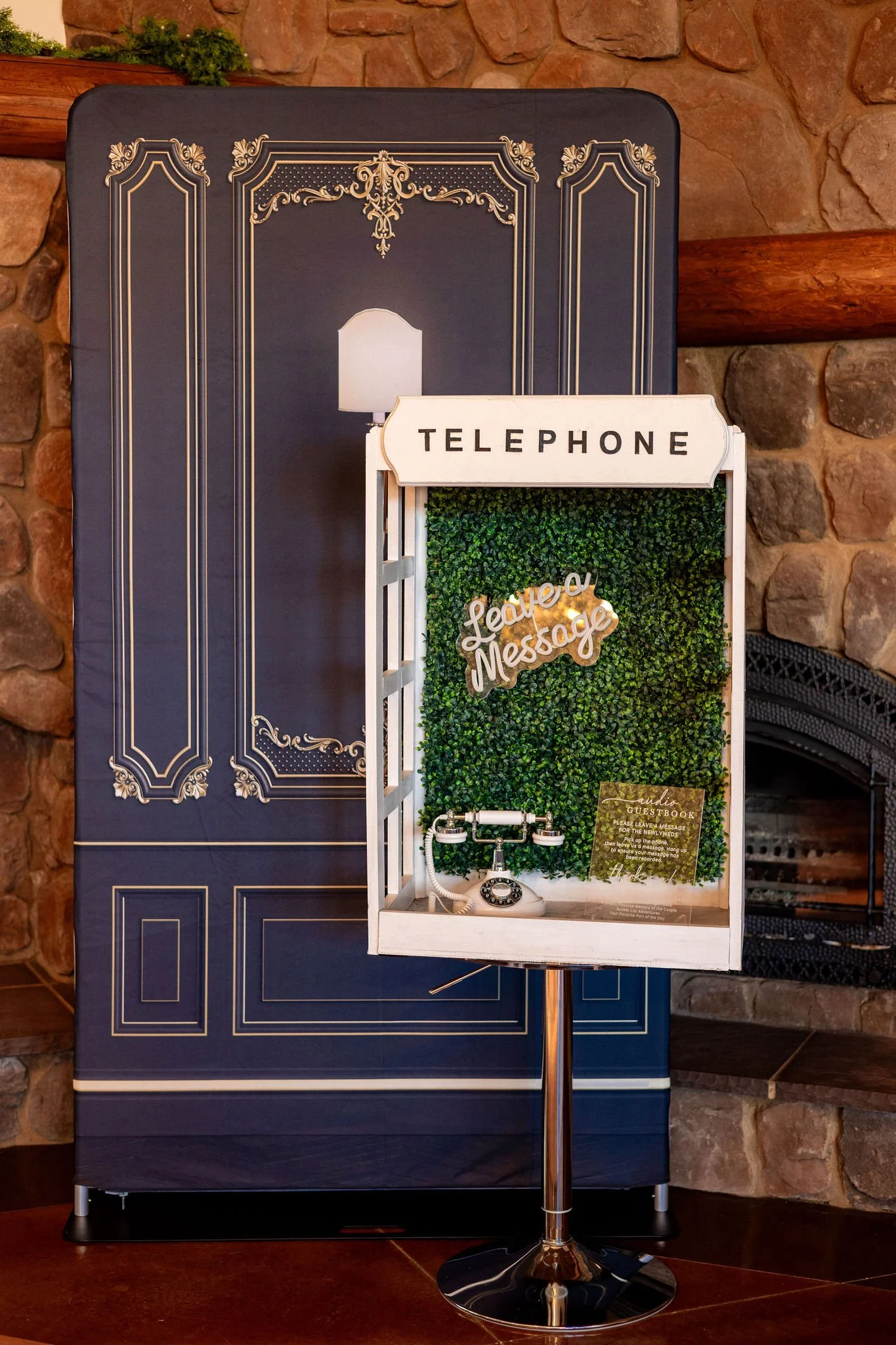 Decorative photo booth with a retro telephone and a green leafy backdrop, labeled 'Picture a Message,' in front of a vintage-style painted wall and stone fireplace.