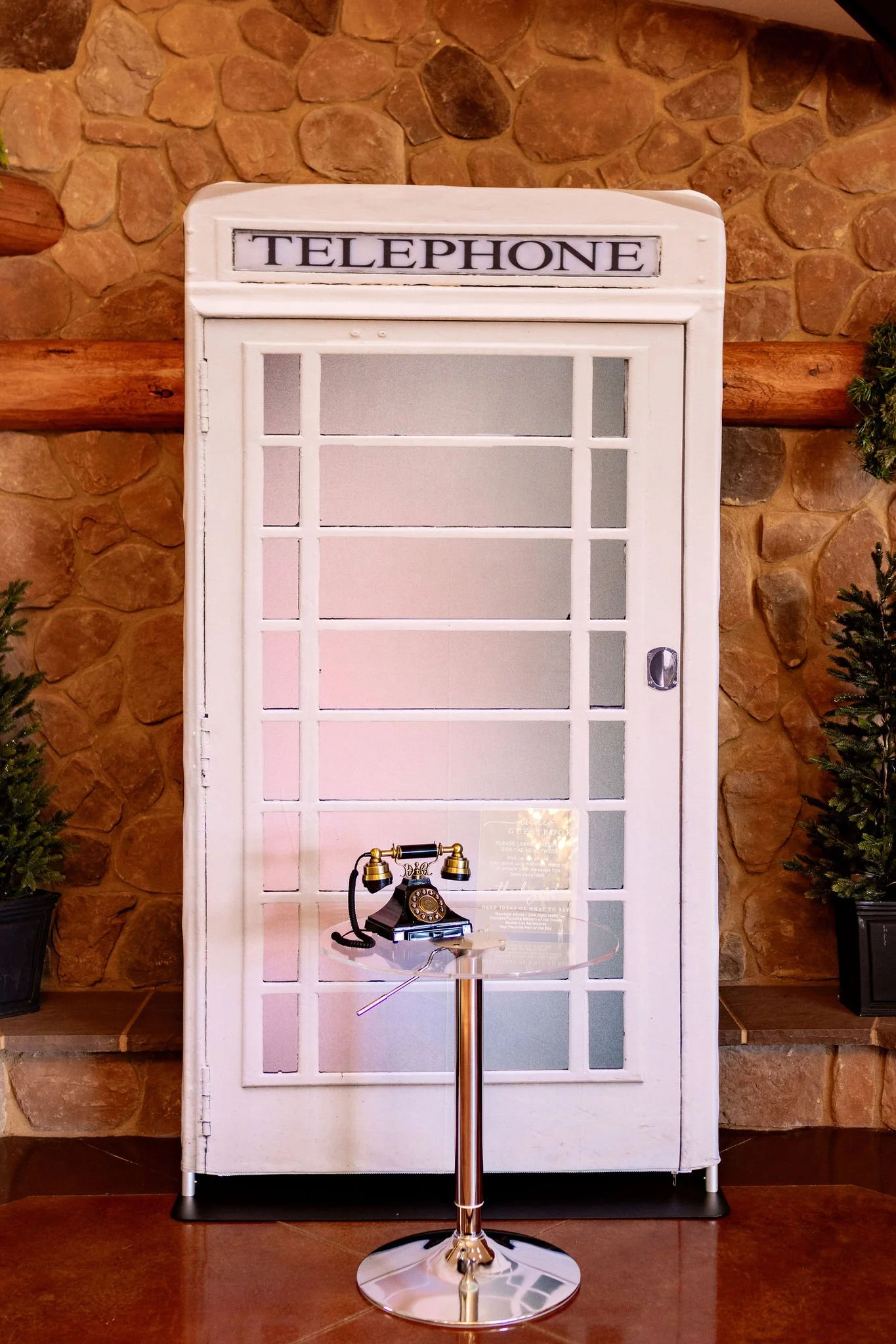 A vintage telephone booth with a black rotary phone on a glass table in front of it, set against a stone wall background.