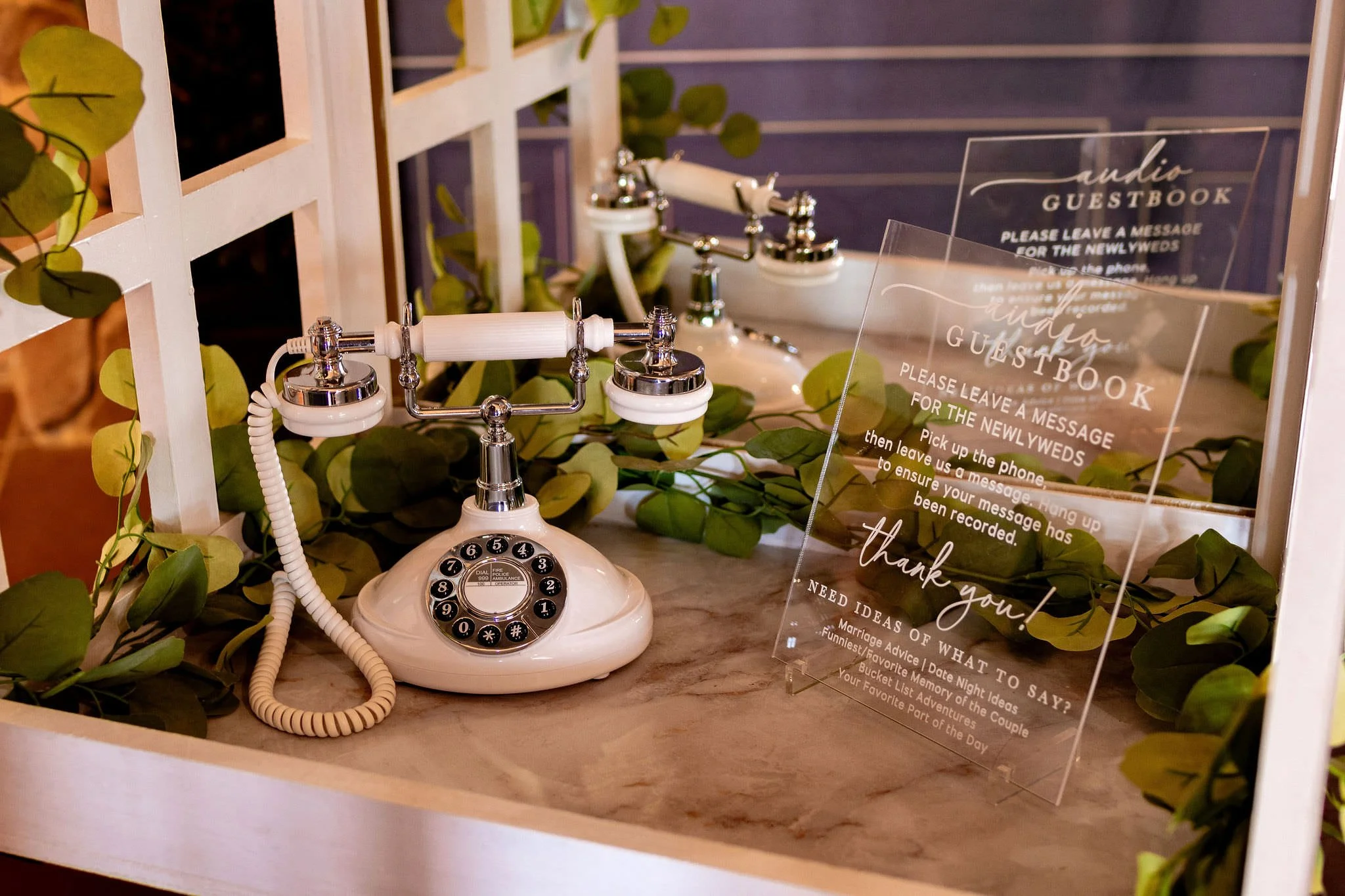 A vintage white rotary telephone with a coiled cord is placed on a marble countertop, surrounded by green leafy vines. A clear acrylic sign is positioned to the right, providing instructions for leaving messages at a guestbook station, with text including "please leave a message for the newlyweds" and "thank you!" Next to the phone, there's a mirror reflecting part of the setup.