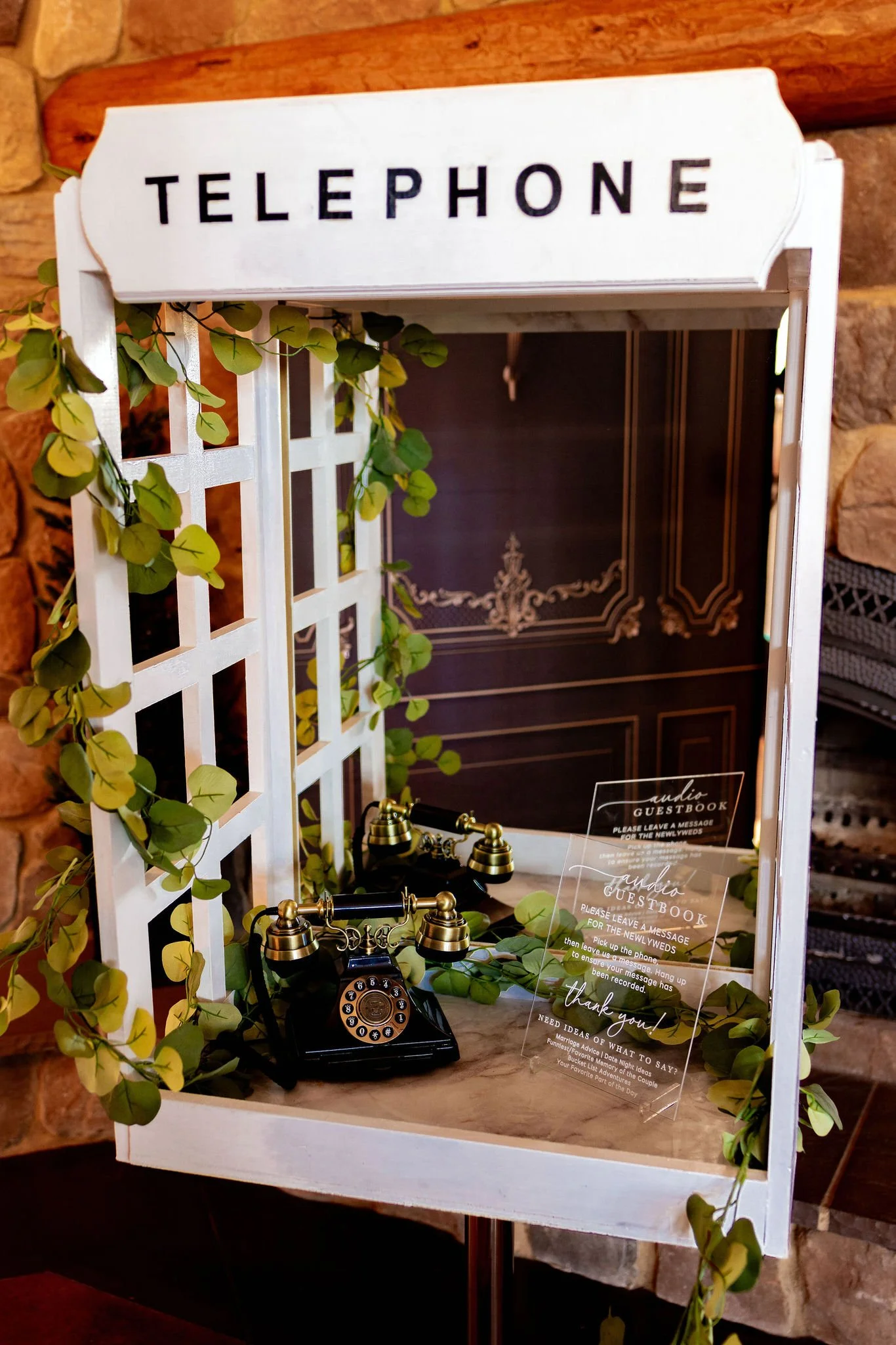Decorative vintage telephone with a sign reading 'Telephone', placed inside a rustic white frame decorated with green foliage, on a wooden surface. A guestbook and a thank you note are also visible.