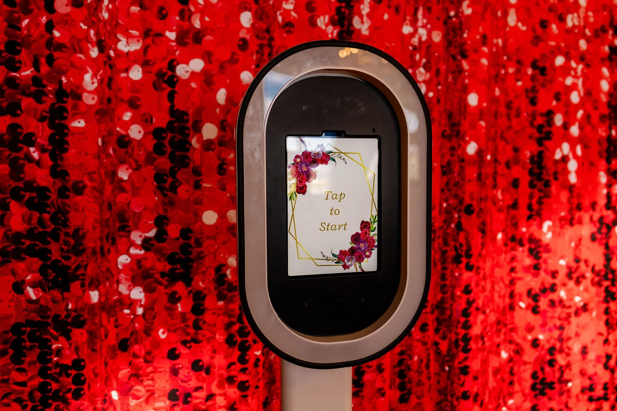 A touchless interaction kiosk with a screen displaying the words 'Tap to Start' surrounded by a floral decoration, set against a red sequined backdrop.