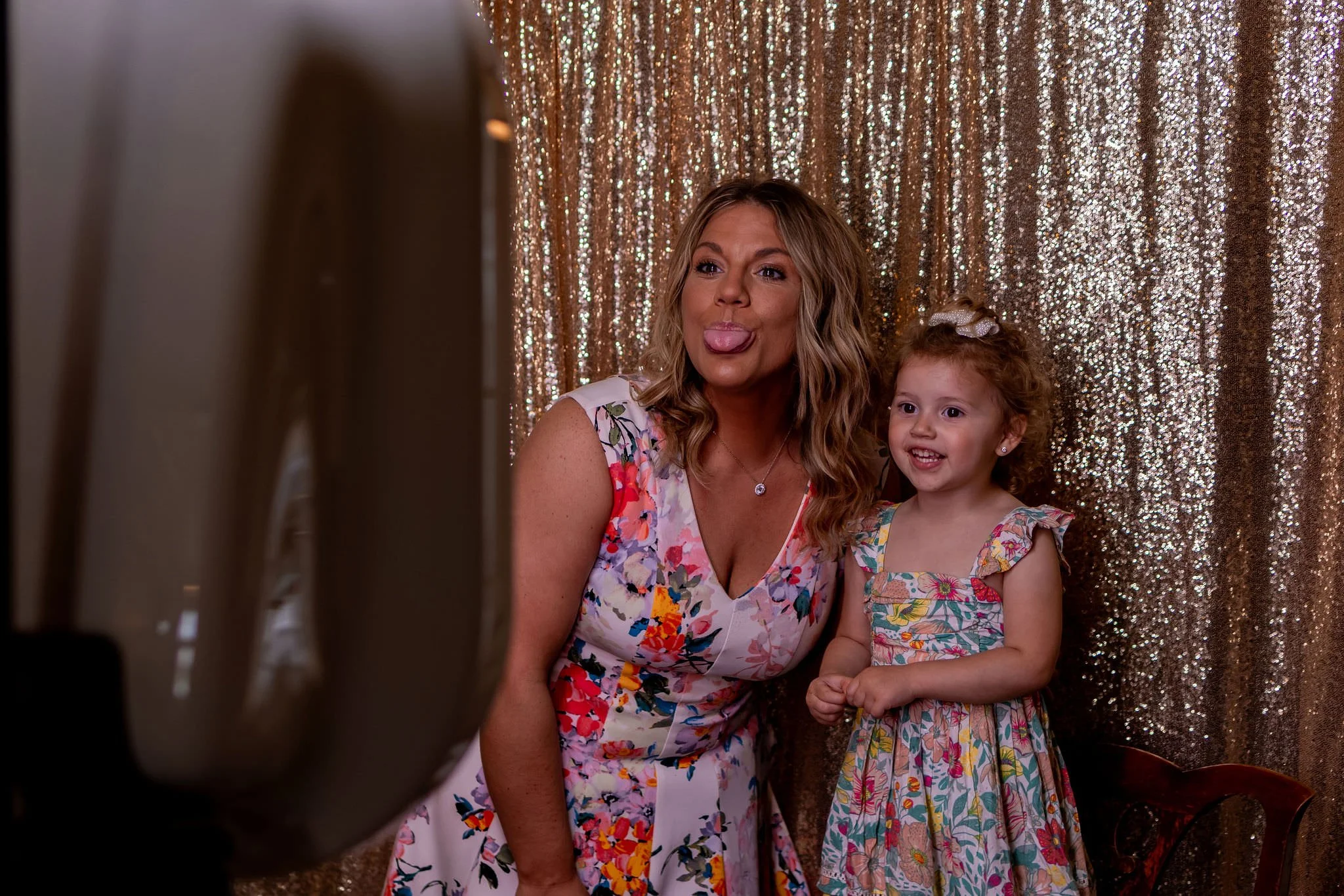 A woman and a young girl taking a selfie together against a sparkly, gold-colored curtain background. They are both wearing colorful dresses, and the woman is sticking out her tongue while the girl is smiling.