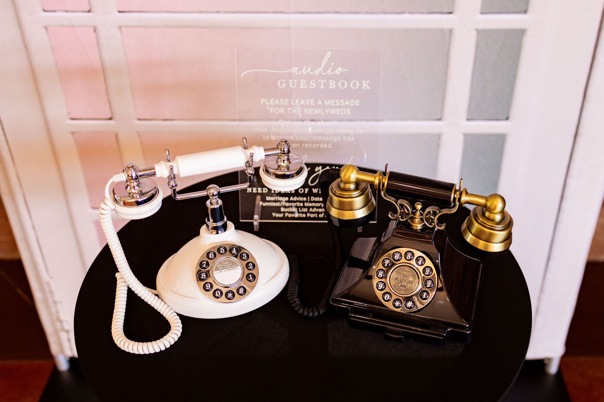 Two vintage rotary dial phones, one white and one black, are placed on a dark surface. A clear acrylic sign with white text about a guestbook is behind them. The background features a white-paneled wall.
