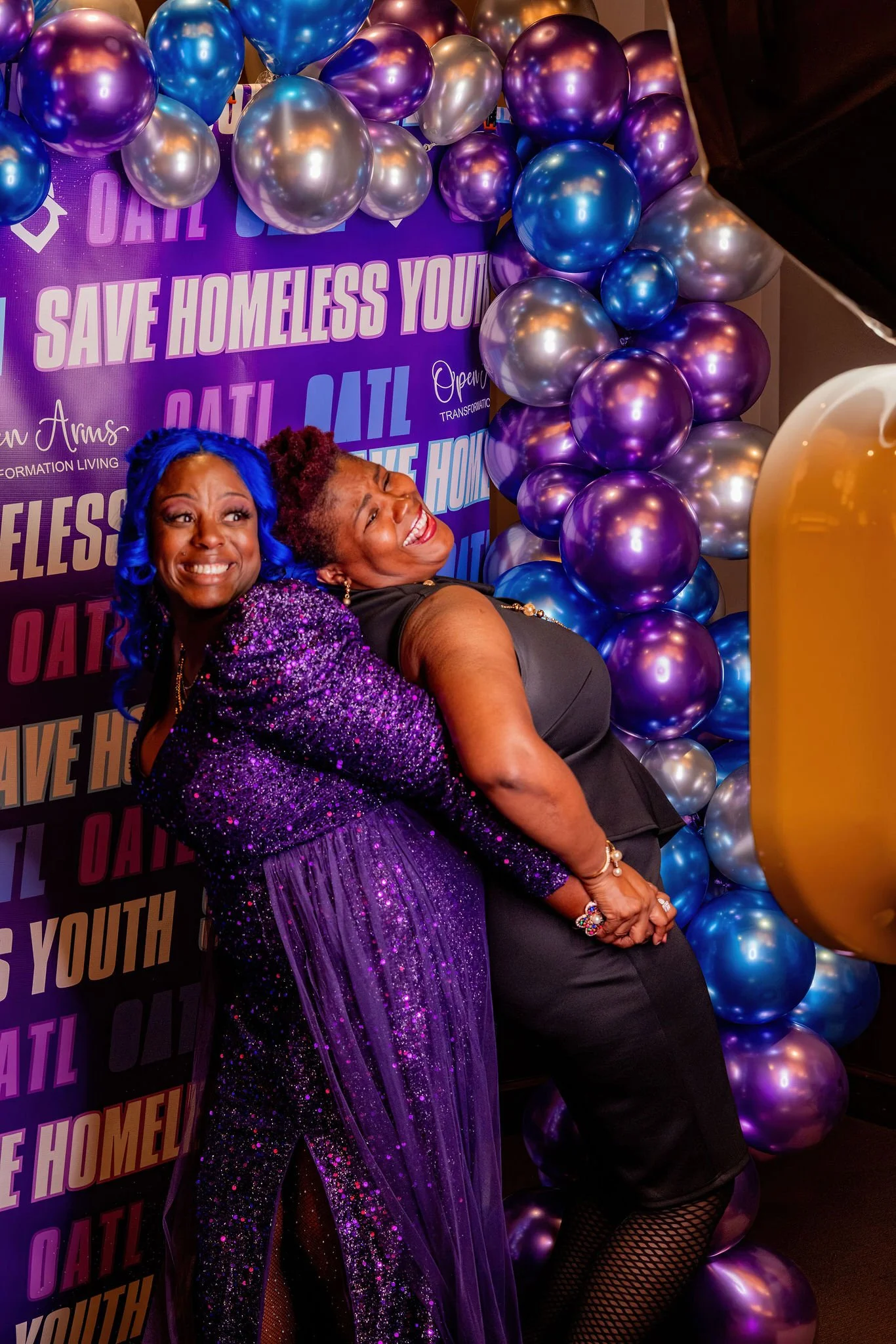 Two women smiling and hugging in front of a backdrop with balloons and text supporting homeless youth.