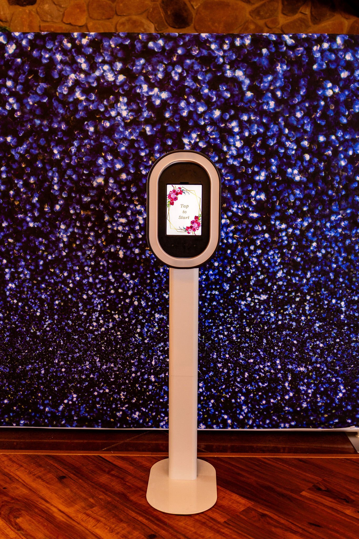 A digital kiosk with a small screen that says 'Tap to Start' is positioned in front of a large floral display in shades of purple and blue, with a brick wall above. The stand is white and placed on a wooden floor.