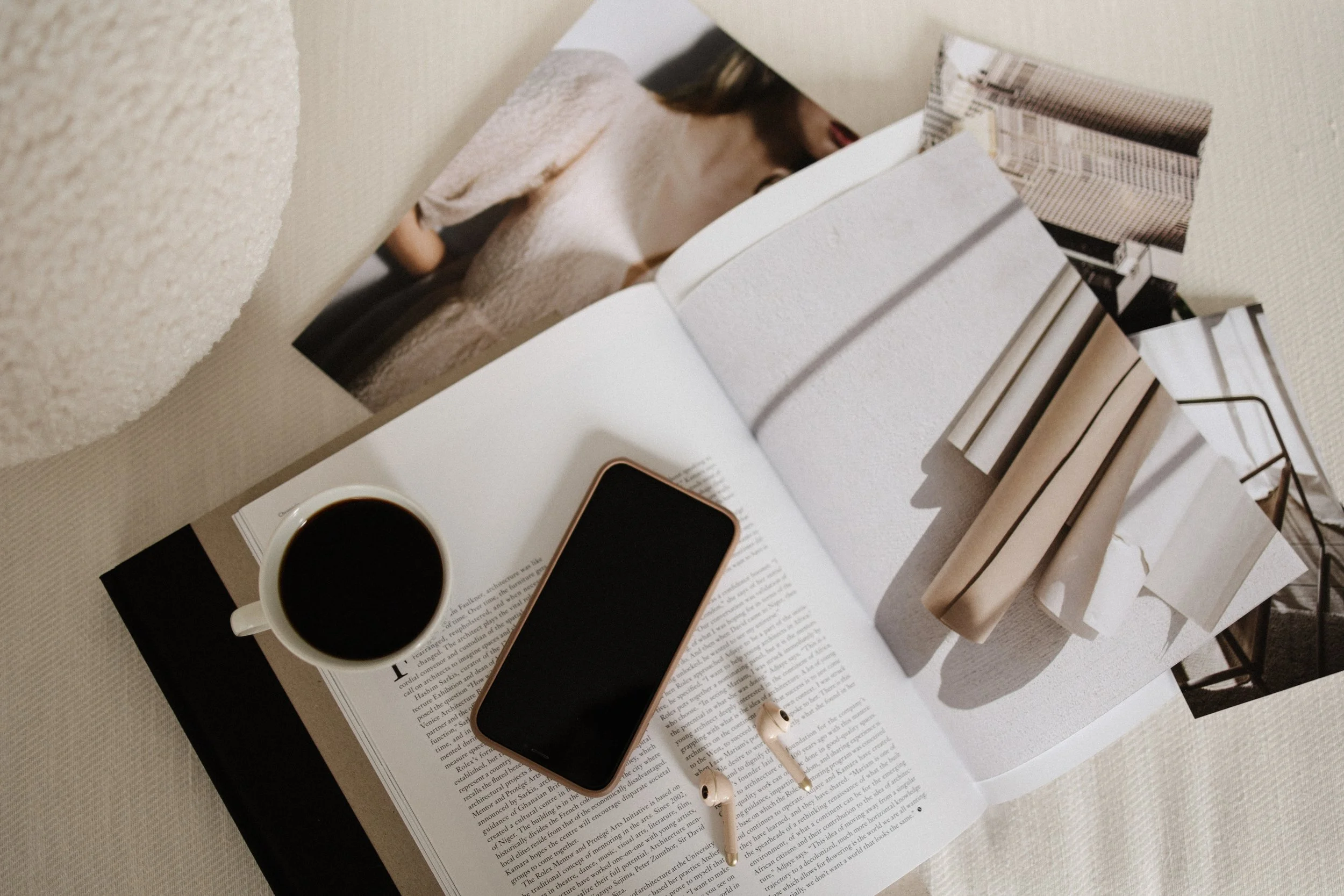 An open magazine with a cup of coffee, a smartphone, and earbuds on top, placed on a textured beige surface with additional photos or pages partially folded underneath.