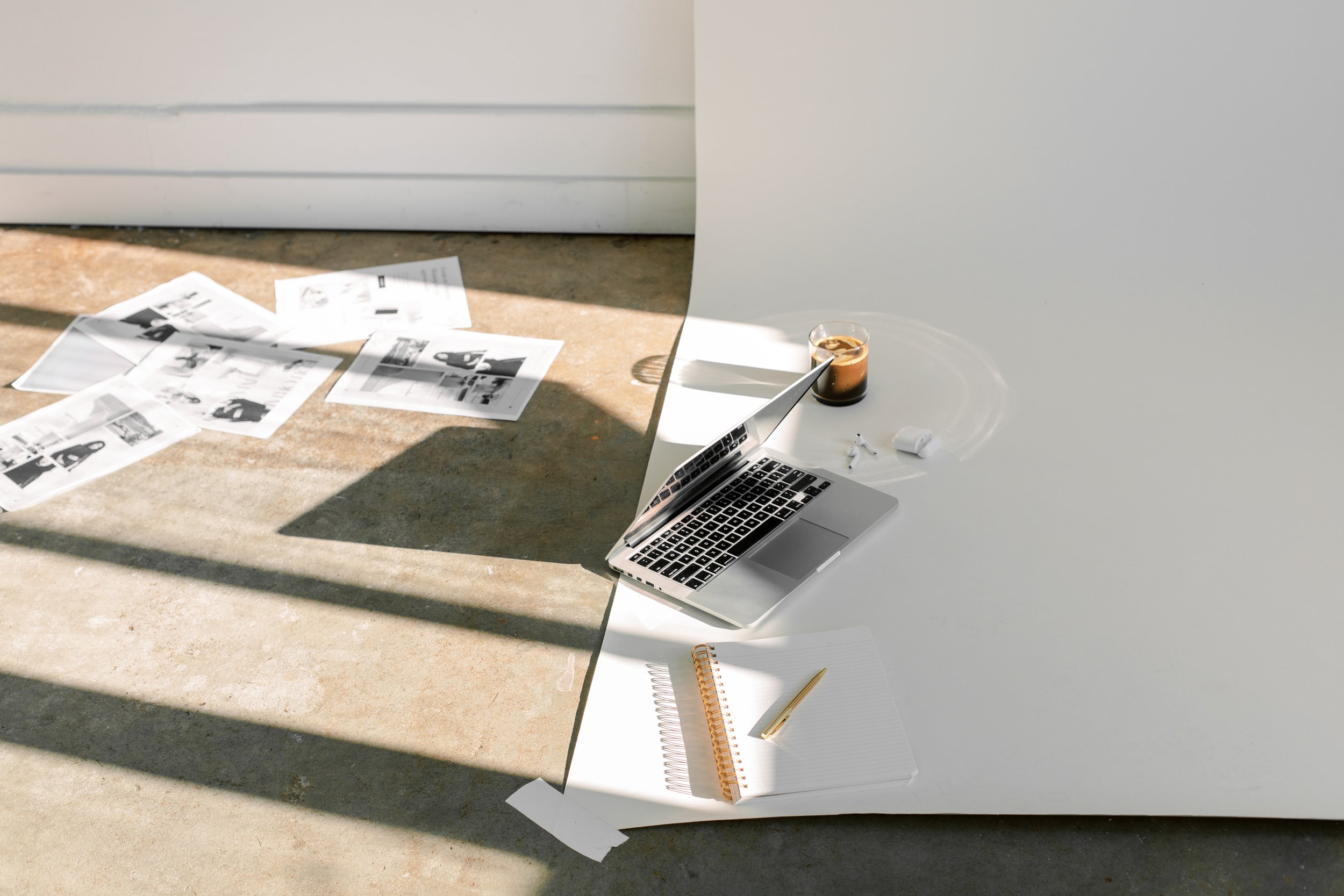 A desk setup with a laptop, a glass of iced coffee, earbuds, a notebook with a pen, and scattered printed papers on a concrete floor, with shadows from sunlight.