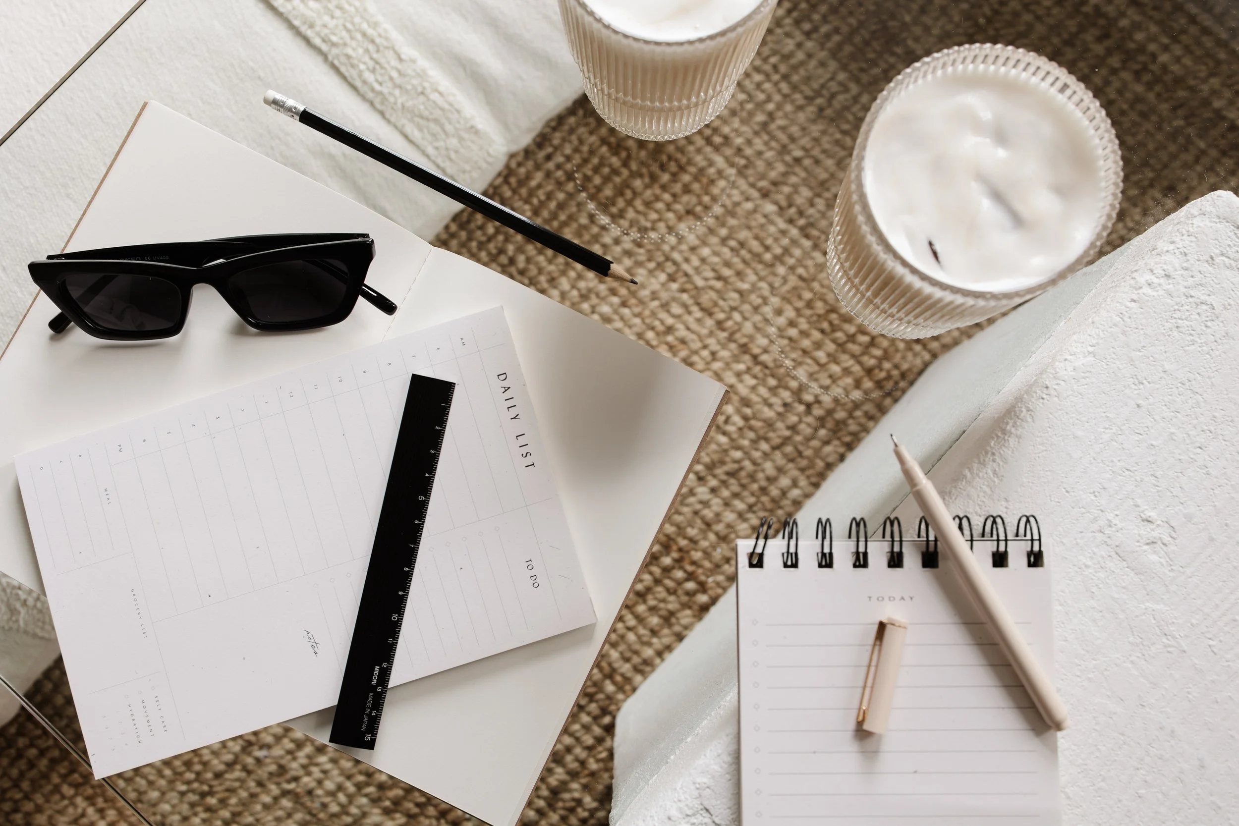 A desk setup with sunglasses, a ruler, a pencil, a spiral notepad, two glasses of iced coffee, and a white planner or notebook, with a textured surface underneath.