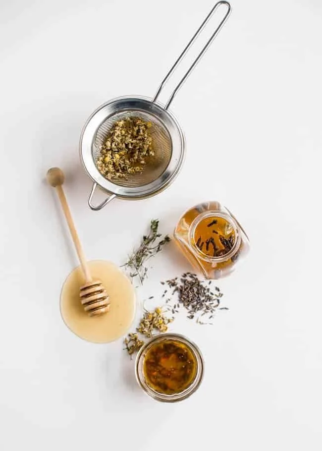 A stovetop strainer with dried chamomile flowers, a honey dipper with honey, a small jar of honey, a jar of tea with herbs, and loose herbs scattered on a white surface.