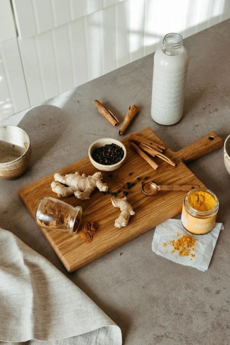 A wooden cutting board with ginger root, black peppercorns, turmeric powder, cinnamon sticks, and a small bowl of black pepper. There is a jar of turmeric powder, a spoon, a piece of paper, an upside-down glass jar, and a bottle of milk or cream on a