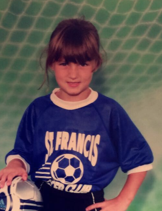 Child girl with brown hair and bangs, wearing a blue sports jersey with 'SF FRANCS' and a soccer ball logo, standing in front of a green net background, holding a soccer ball.