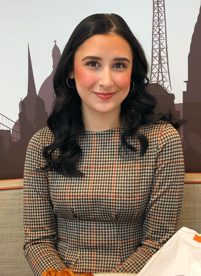 Young woman with dark hair, wearing a houndstooth patterned top, sitting at a table with a city skyline mural in the background.