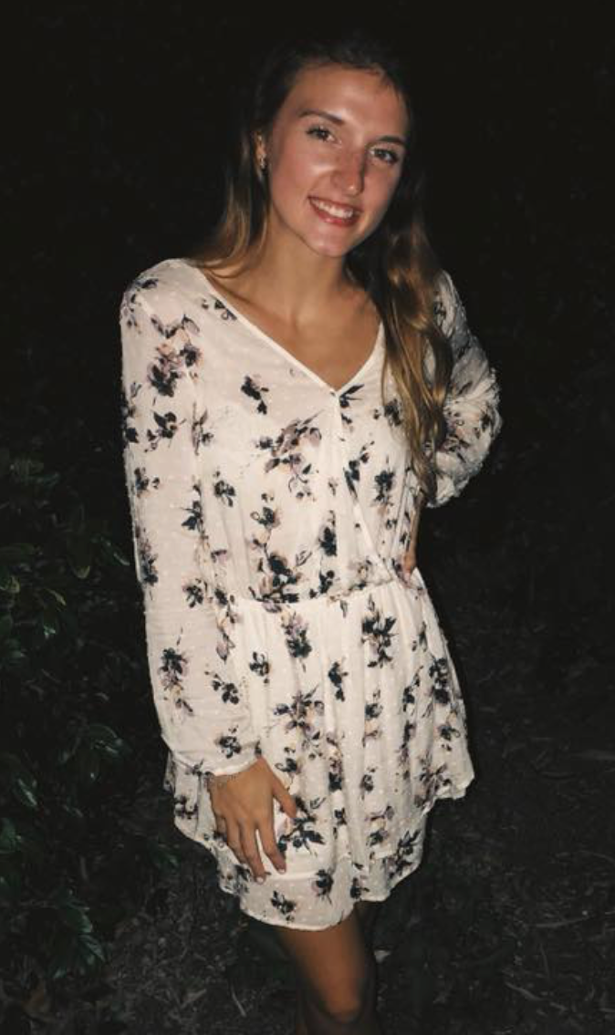 A young woman smiling, wearing a white floral dress with long sleeves, standing outdoors at night.