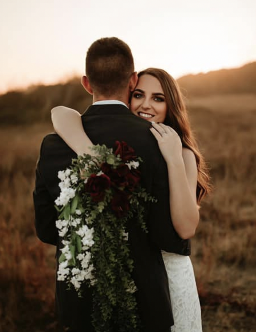 A newlywed couple embracing outdoors at sunset. The bride is smiling while hugging the groom's shoulder, holding a cascading bouquet of red and white flowers.